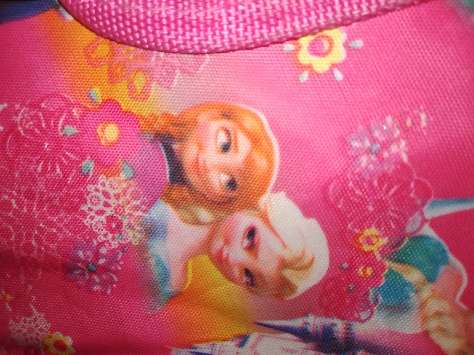 The image is a close-up of a pink fabric with a colorful print. The print features stylized cartoon characters, possibly from a children's show, with large eyes and smiling faces. There are also floral patterns in yellow and white scattered across the fabric. The texture of the fabric is visible, showing a woven pattern. The lighting is bright, creating some glare on the surface. The image does not contain any information or visual cues that would allow for identification of a specific location in Santa Ana, Venezuela.