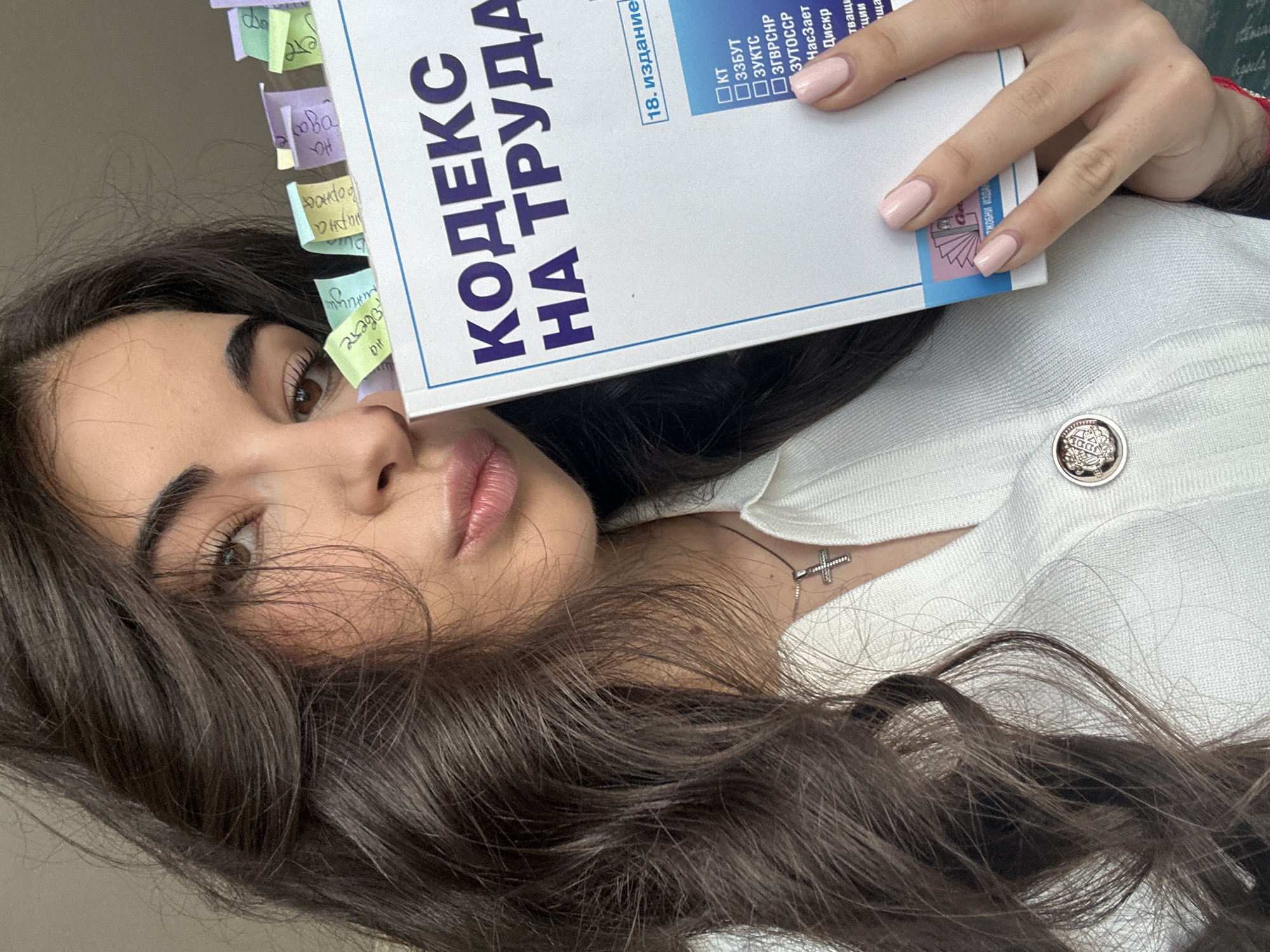 A young woman with long, dark, wavy hair is holding a book titled "KODEKC HA TRUDA" (Labor Code) in front of her face. Her right hand is visible, holding the book, and she has light pink nail polish. The book has several colorful sticky notes attached to its pages. The woman is wearing a white, ribbed top with a silver cross necklace. The background is blurred and appears to be indoors, with soft lighting. The scene suggests the woman is studying or reviewing the book. There is no information in the image to suggest the location is Sofia, Bulgaria.
