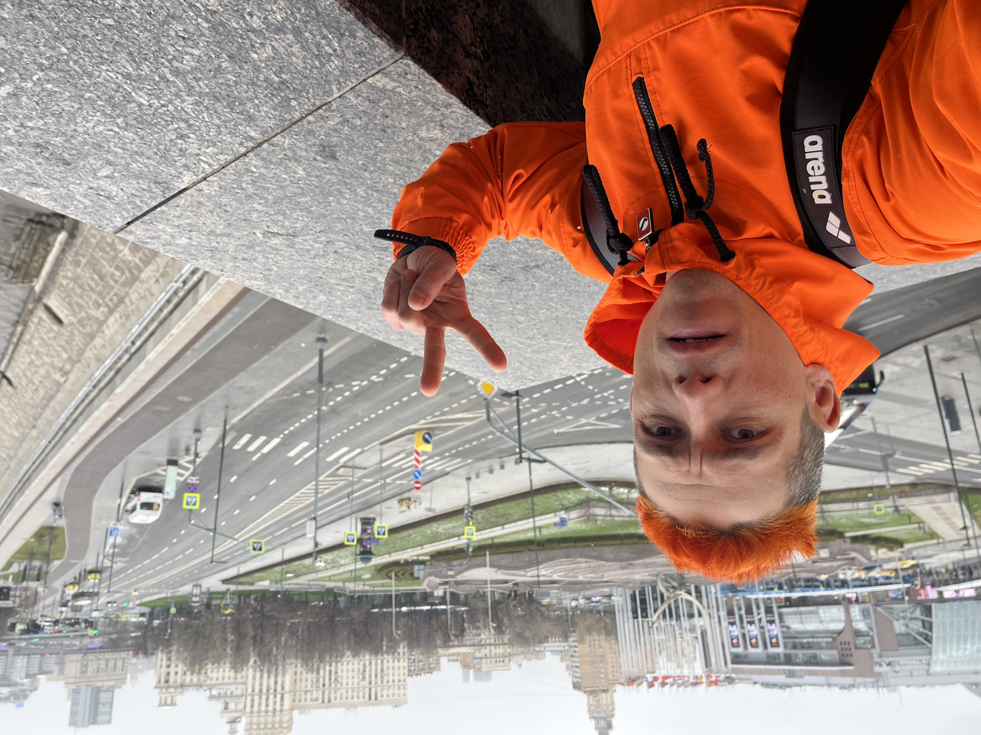 A medium shot shows a person with orange hair, wearing an orange jacket, and a black "arena" strap. The person is holding up two fingers in a "peace" sign. They are standing in front of a wide city street with vehicles and pedestrian crosswalks. The background reveals buildings typical of Moscow, Russia.