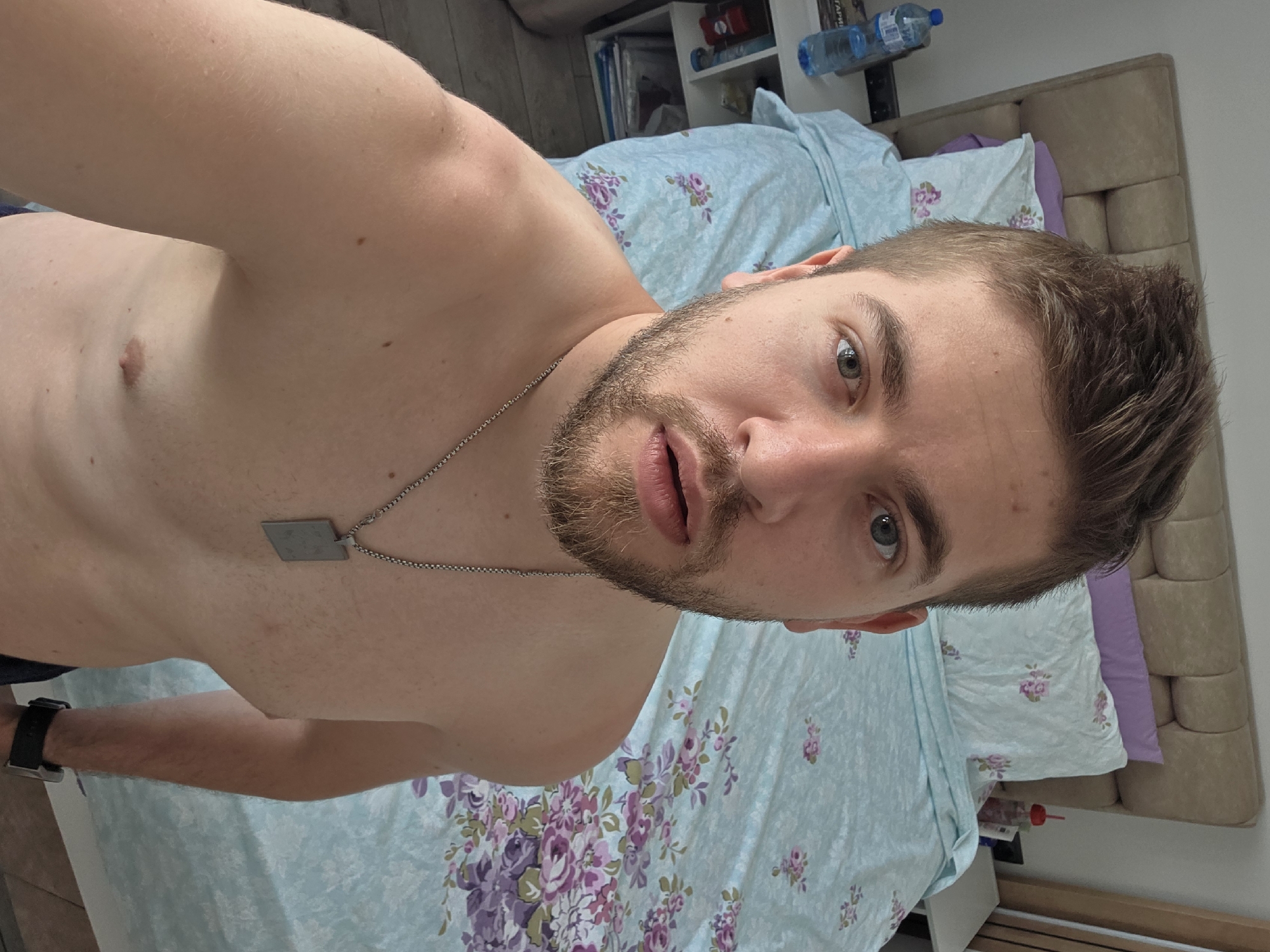 The scene is an indoor setting, identified as a bedroom in the city of Yagodovo, Bulgaria. A Caucasian male, estimated to be in his 20s or early 30s, is captured in a close-up, upward-angled photograph, appearing to be a selfie. He has short, styled light brown hair, light-colored eyes, and a short beard. His mouth is slightly open, and he maintains direct eye contact with the camera. The man is bare-chested, wearing a silver chain with a rectangular dog tag-style pendant around his neck and a dark-colored smartwatch on his left wrist. Visible skin exhibits several small moles or freckles and some minor redness on the shoulders.
In the background, a bed with a light brown upholstered headboard is partially visible. The bed is covered with light blue bedding adorned with a pattern of pink and purple floral clusters. One plain purple pillow and one matching floral pillow are positioned against the headboard. To the left of the bed, a white multi-tiered nightstand holds a clear plastic water bottle with a blue cap and a book or magazine displaying Cyrillic text, with a visible fragment suggesting "БЪЛГАРИЯ" (Bulgaria). To the right of the bed, a white nightstand holds a red-capped bottle or cup. A light-colored wall is visible on the left, and a wooden vertical panel or door frame is present on the far right.