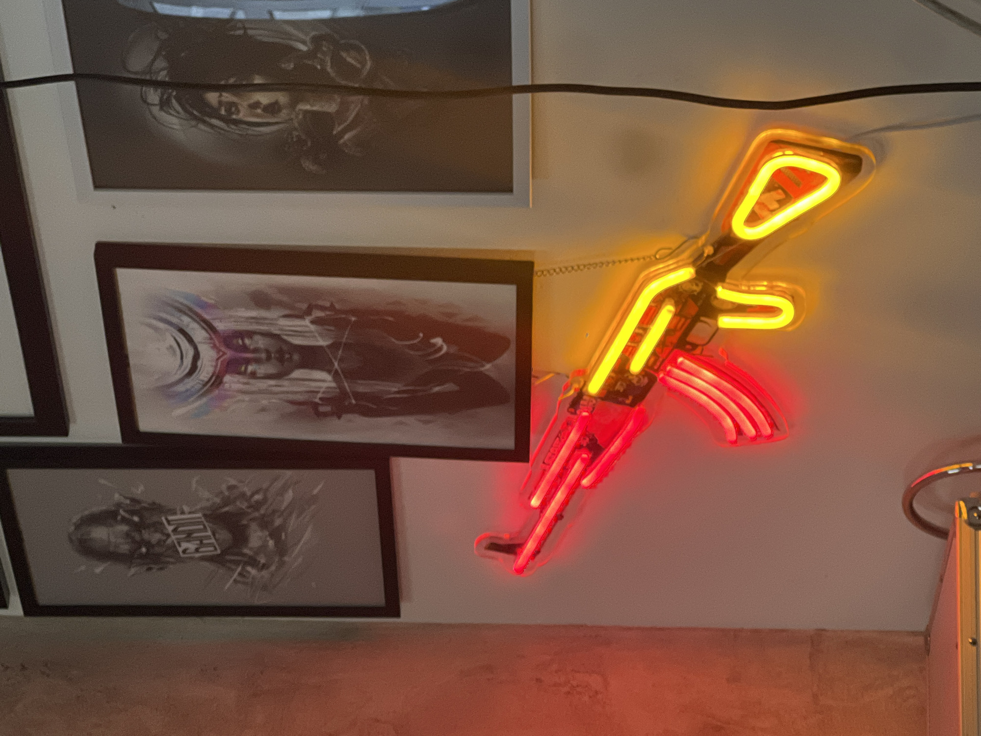 The image shows a room with three framed pictures hanging on the wall to the left and an illuminated neon sign of a rifle on the right. The neon sign has a yellow tube for the barrel and stock, and a red tube for the magazine. The pictures are framed in black and white frames. The wall is painted white and the floor is tiled in a neutral color. There is a power cord running across the top of the image. Based on the style of the art, the decor, and the presence of Cyrillic text on one of the pictures, it is possible this scene is located in Sofia, Bulgaria.