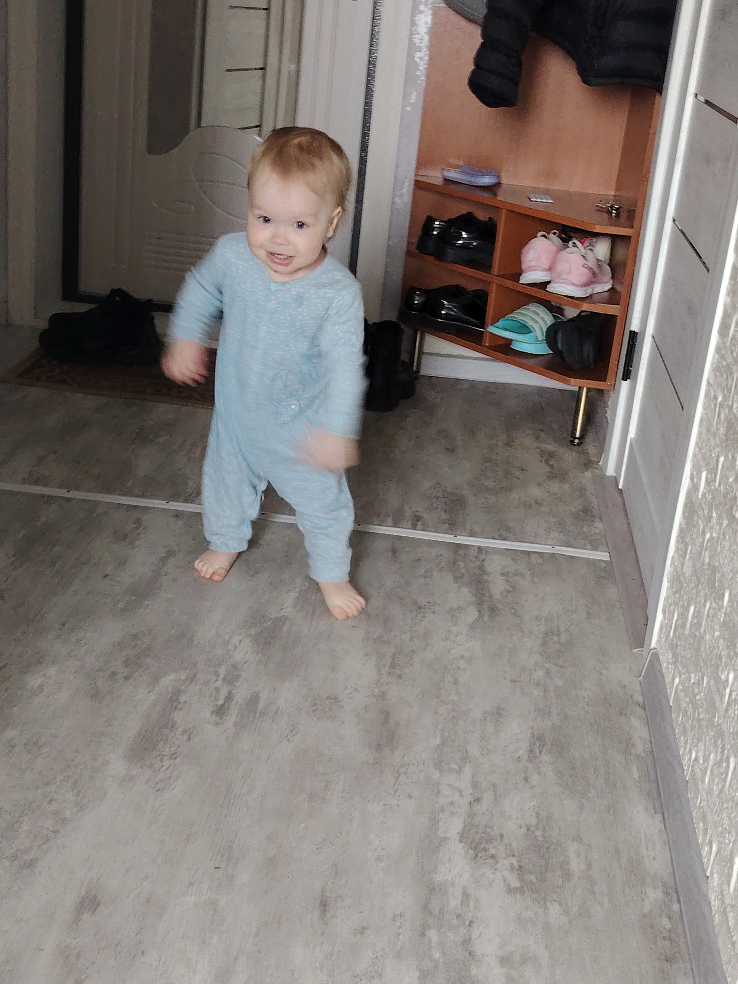 A child stands in a doorway inside a building. The child has blonde hair and is wearing a light blue onesie. The child is smiling. In the background, there is a shoe rack with shoes and a jacket hanging above it. The location is in a building in the city of Chaikovsky, Russia. The floor is gray. There is a white door in the background.