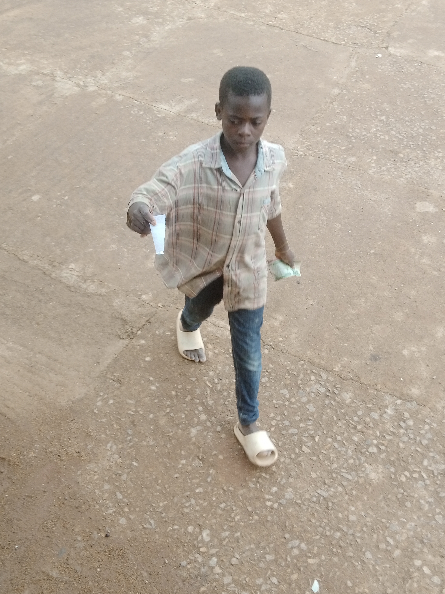 A young Black boy walks on a paved surface. He is wearing a plaid button-down shirt, blue jeans, and light-colored slide sandals. In his right hand, he holds a small white object, possibly paper. In his left hand, he holds a green bill, which appears to be a banknote. The boy is looking down and to his left. The ground is a mix of dirt and small stones. The lighting suggests it is daytime, but the weather is not discernible. The scene appears to be outdoors, possibly a street or pathway.