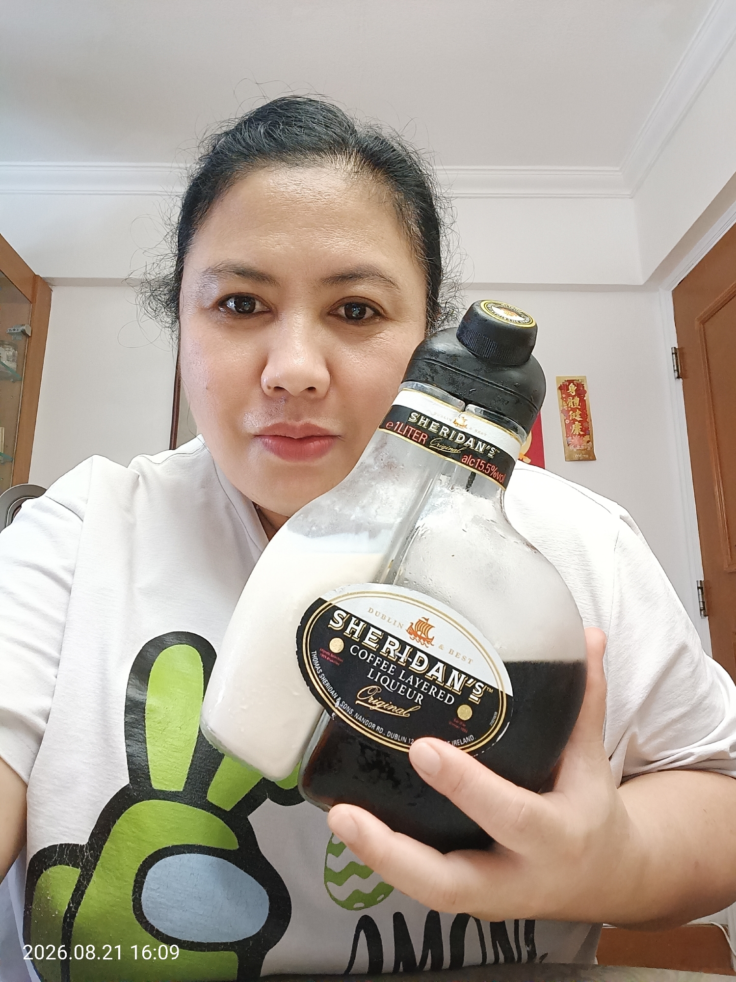 A woman is holding a bottle of Sheridan's coffee layered liqueur. The bottle has two compartments, one filled with a white creamy liquid and the other with a dark coffee liqueur. The woman is wearing a white t-shirt with a green cartoon character printed on it. She has dark hair and is looking directly at the camera. The background shows a white wall, a wooden door, and a cabinet. There is also a Chinese scroll with red characters hanging on the wall. A timestamp in the lower left corner indicates the date and time as 2026.08.21 16:09. The image does not provide any specific information about the location within Hong Kong, China.
