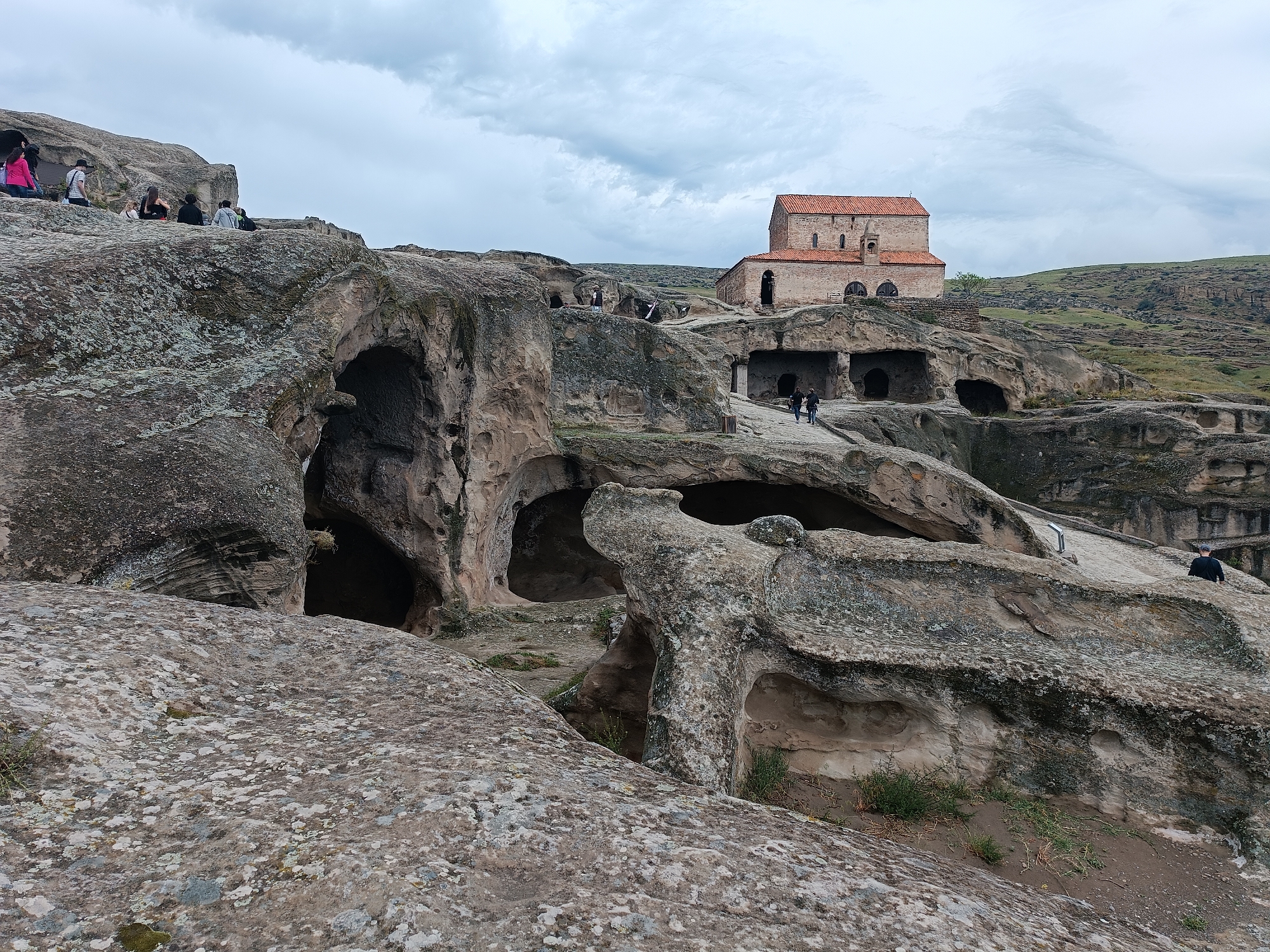 The image depicts the ancient cave city of Uplistsikhe in Georgia. In the foreground, large, weathered rock formations with visible cave entrances dominate the landscape. Patches of green vegetation sprout from the rocky ground. In the middle ground, several people are scattered across the rock formations, some sitting and others walking. A stone pathway winds through the area. Further in the background, a stone church with a red-tiled roof stands prominently on a hill. The sky is overcast with clouds.