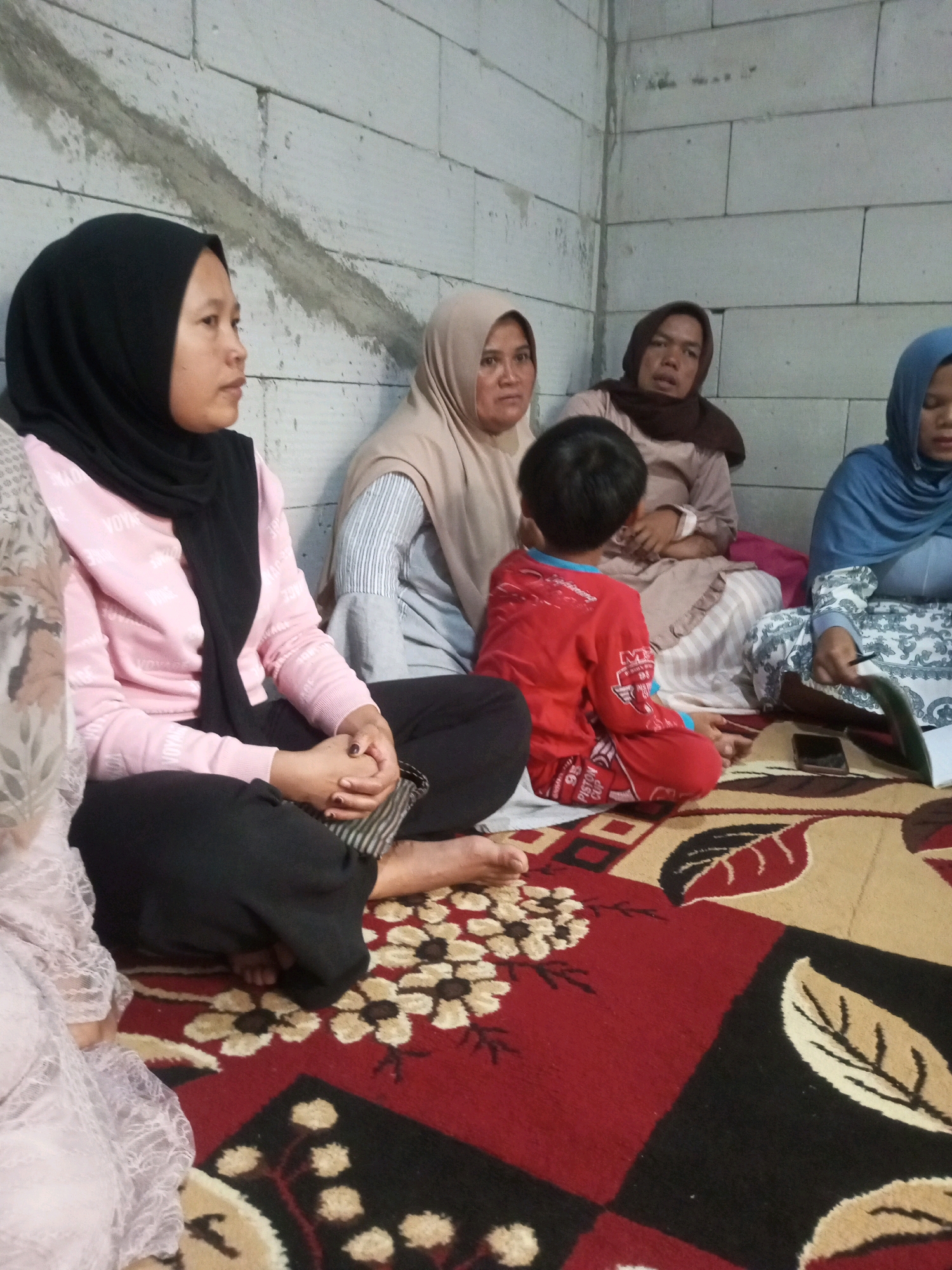 Five people are seated on a patterned rug in what appears to be an indoor setting with a brick wall in the background. On the left, a woman wearing a black hijab and a pink sweater is looking to the side. Next to her, a woman in a light brown hijab is also looking to the side. In the center, a young boy in a red shirt is facing away from the camera. To his right, a woman in a dark brown hijab is looking forward. On the far right, a woman in a blue hijab is looking down at a piece of paper and a cell phone. The rug has a prominent floral and geometric pattern in red, black, and beige. The setting is consistent with a residential interior in Andongsili, Indonesia, likely a living room or communal space.