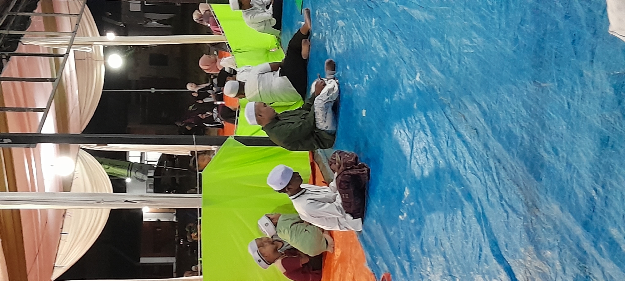 In Bendungan, Indonesia, a group of people are gathered outdoors under a tent structure. The ground is covered with blue and orange tarpaulins. Several individuals, predominantly male, are seated on the tarpaulins. Some are wearing white skullcaps and traditional Islamic attire. A few younger individuals, also in skullcaps and casual clothing, are seated together. One man in a green shirt is looking down at his phone. Another man in a white shirt is reading from a book or tablet. The scene appears to be a religious gathering or event taking place in the evening, indicated by the artificial lighting.