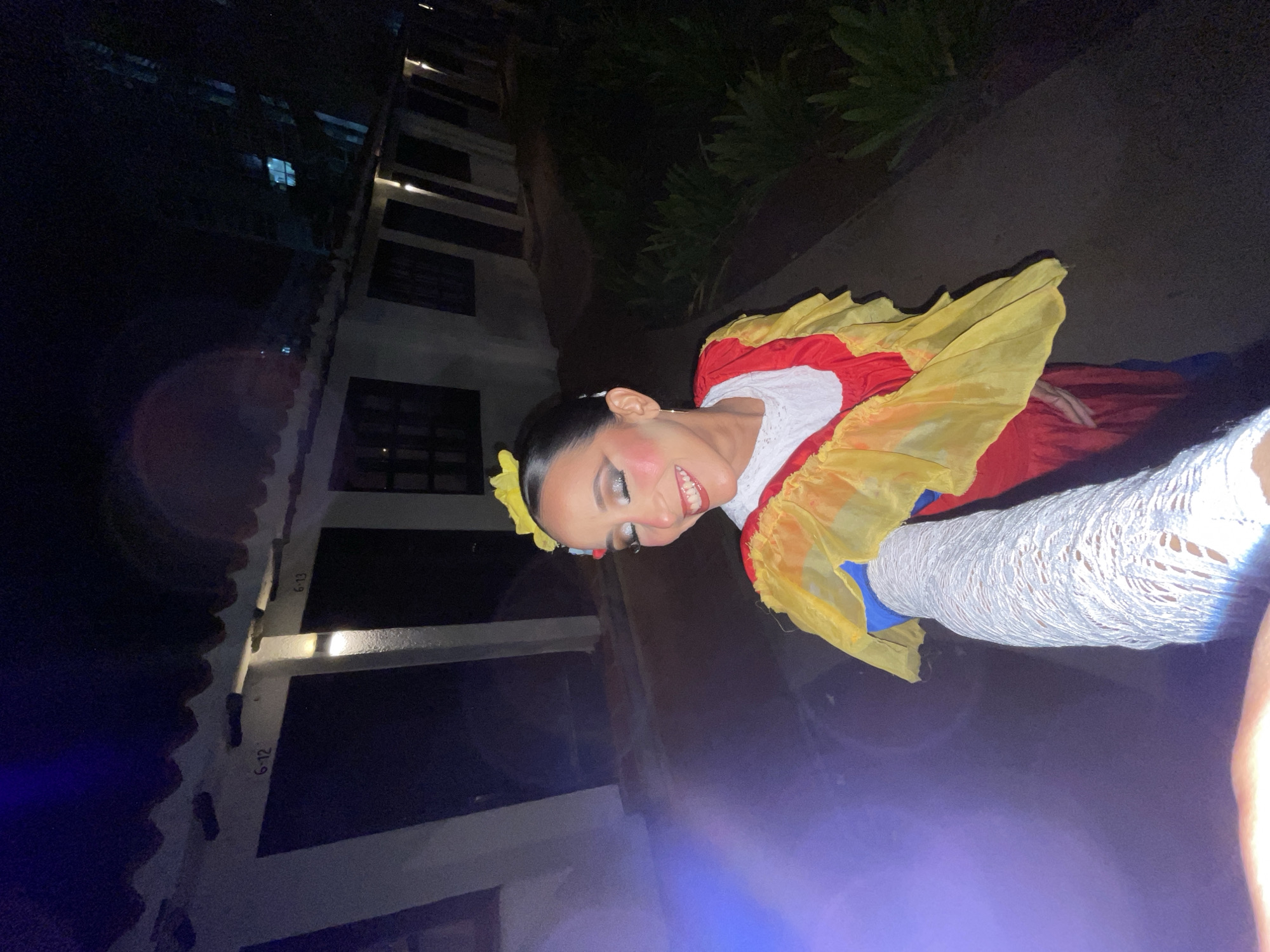 A selfie taken at night shows a woman in traditional Colombian dance attire. She is smiling and looking down and to her left. The costume is red with yellow and blue ruffles. She is wearing white lace sleeves and a yellow flower in her hair. The background appears to be an outdoor walkway in front of buildings labeled "6-12" and "6-13." These buildings are likely part of a hotel or resort complex, possibly near the beach in Porlamar, Venezuela. The lighting is dim, with artificial lights illuminating the walkway and the buildings.
