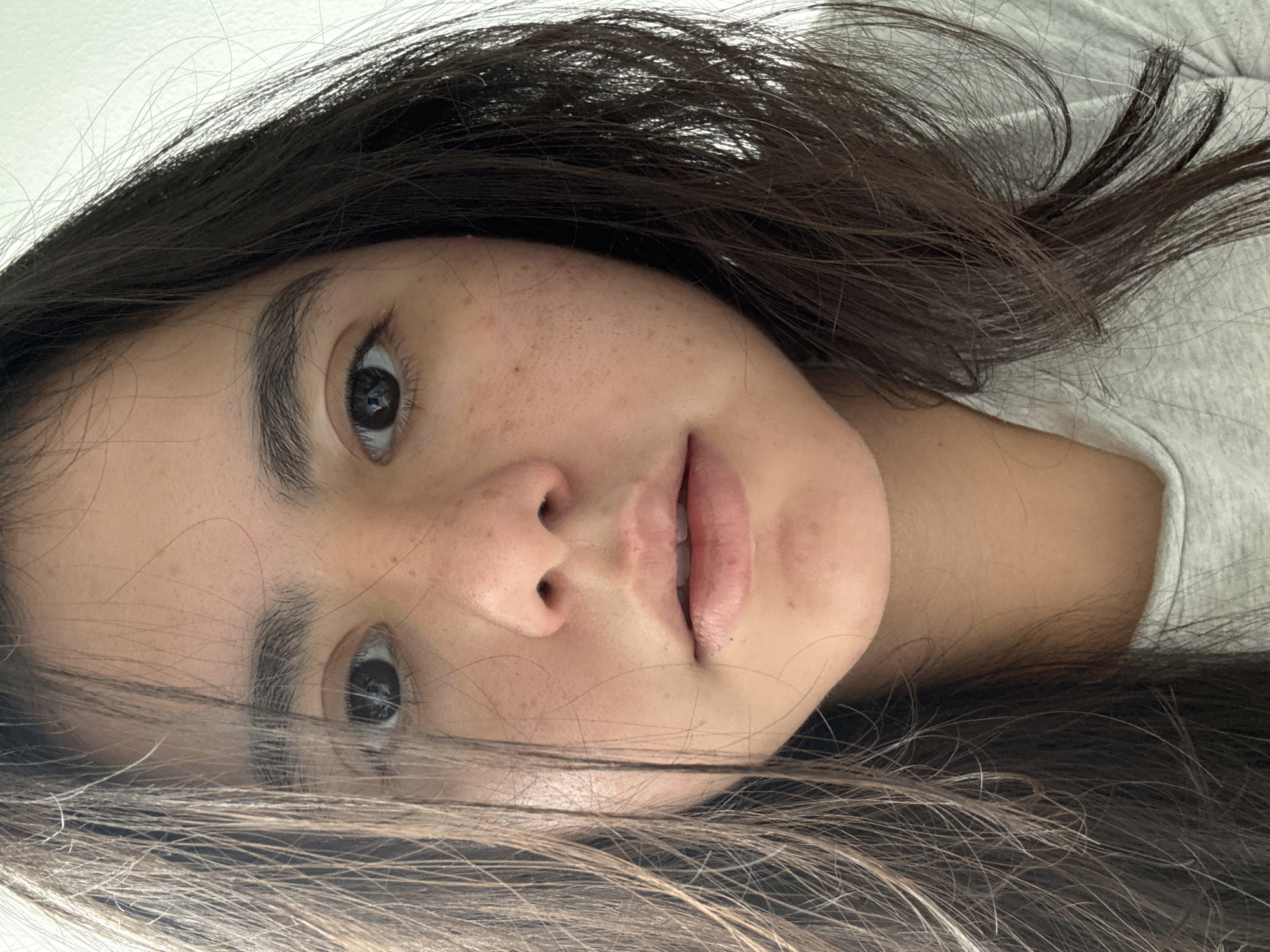 A close-up portrait captures a young woman with dark brown hair, oriented sideways, looking directly towards the viewer. She appears to be lying down, with her head slightly tilted. Her long, dark hair frames her face, with some strands falling across her forehead and shoulders, resting on a light-colored, possibly grey, casual top.
Her dark eyes are open, with visible reflections indicating a soft light source. Her skin is fair, adorned with numerous freckles concentrated across her nose and cheeks. Her lips are naturally pink and slightly parted, revealing a hint of her upper teeth. Her eyebrows are dark and well-defined. The expression on her face is neutral and calm. The background behind her head is a plain, light surface, suggesting an indoor setting. There is no visible text or additional objects in the image.