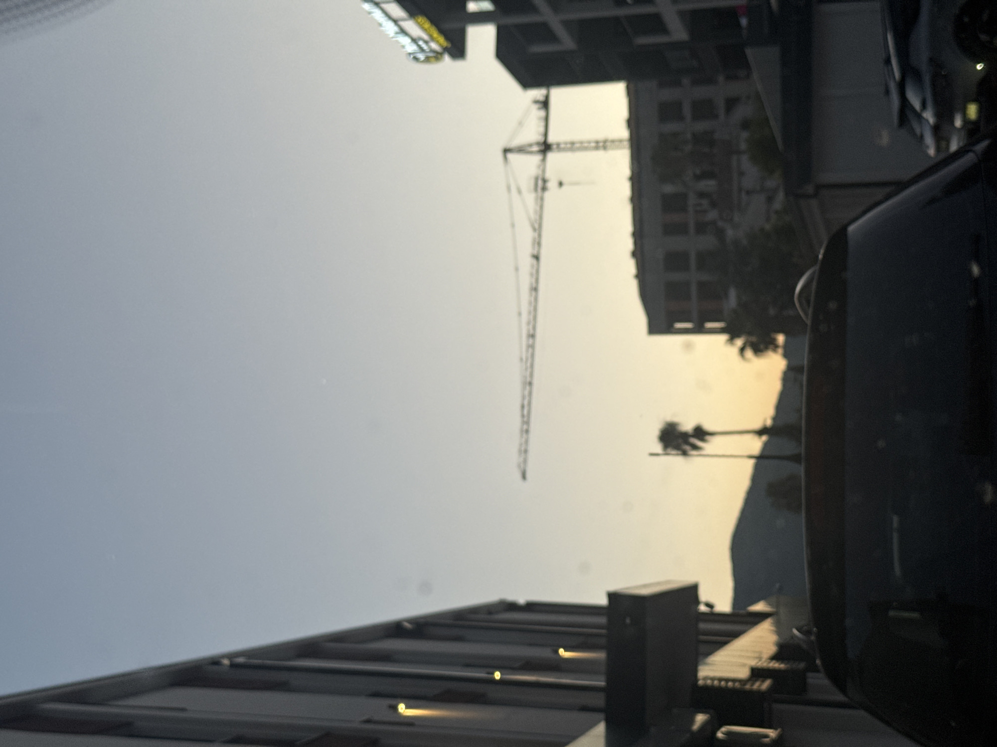 The image depicts a city scene during dusk or dawn. In the foreground, the roof of a dark-colored vehicle is visible. Beyond the vehicle, several buildings of varying heights and architectural styles are present. A prominent construction crane stands in the middle ground, suggesting ongoing development. Palm trees are scattered throughout the scene, hinting at a warm climate. In the background, hills or mountains are visible against the soft glow of the sky. The overall atmosphere is one of urban development combined with natural landscape. There are no people or specific actions clearly discernible in the image. The location is not identifiable as Budva, Montenegro.