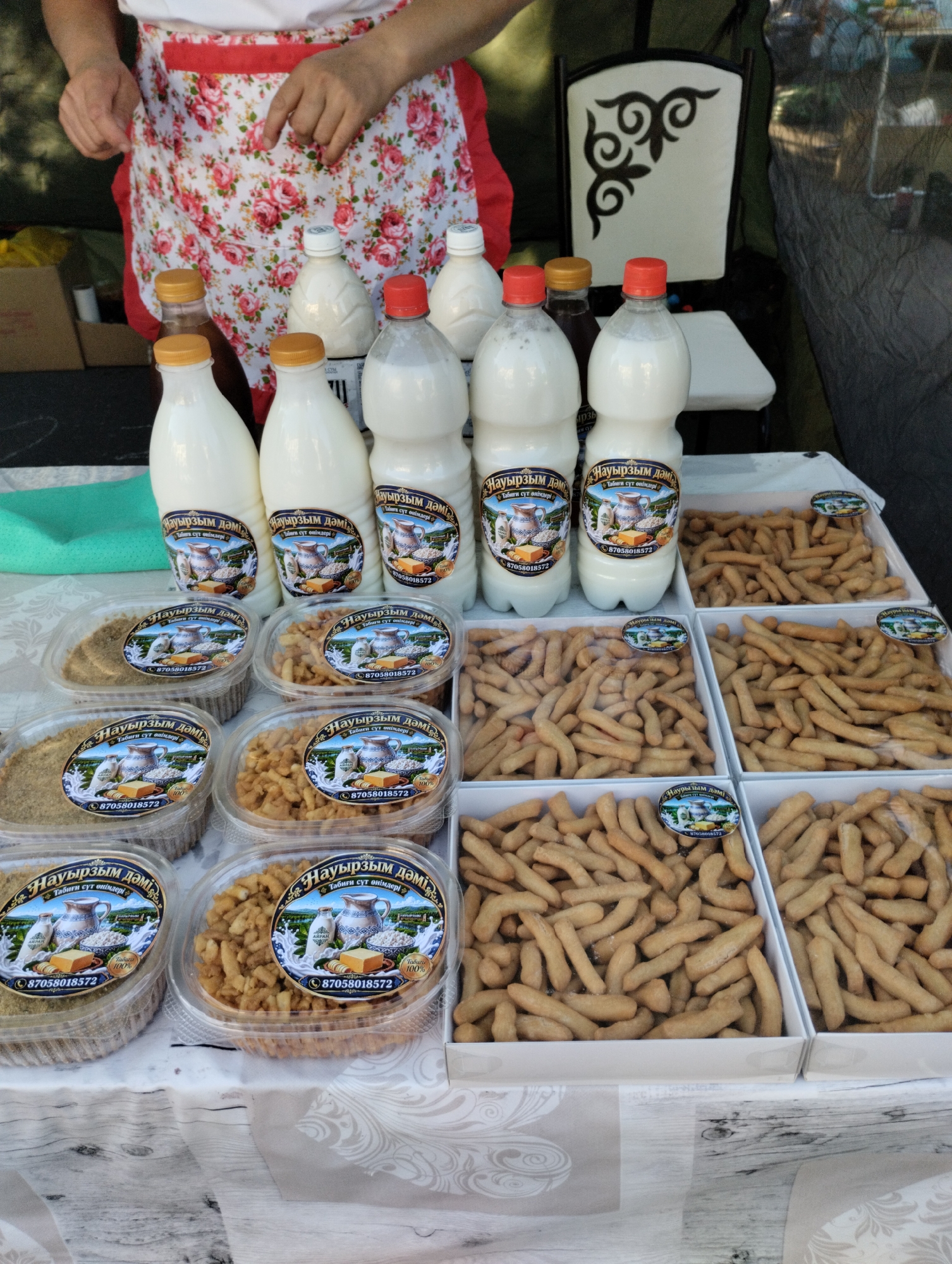 In Kostanay, Kazakhstan, a stall displays an array of food products. In the background, a person wearing a floral apron stands partially visible, with their hands engaged with the items on display. Several clear plastic bottles filled with a white liquid, likely milk or yogurt, are lined up on the table. Each bottle is adorned with a label featuring a pastoral scene, including a jug, cheese, and milk, along with text in a foreign script and a phone number. Alongside the bottles are numerous clear plastic containers filled with a light brown, granular substance. In front of these, rows of white shallow boxes are filled with numerous elongated, golden-brown baked goods, possibly breadsticks or a type of cookie. The stall is set up outdoors, with a glimpse of a dark mesh curtain and a chair with an ornate pattern in the background. The table is covered with a white tablecloth with a subtle floral pattern.