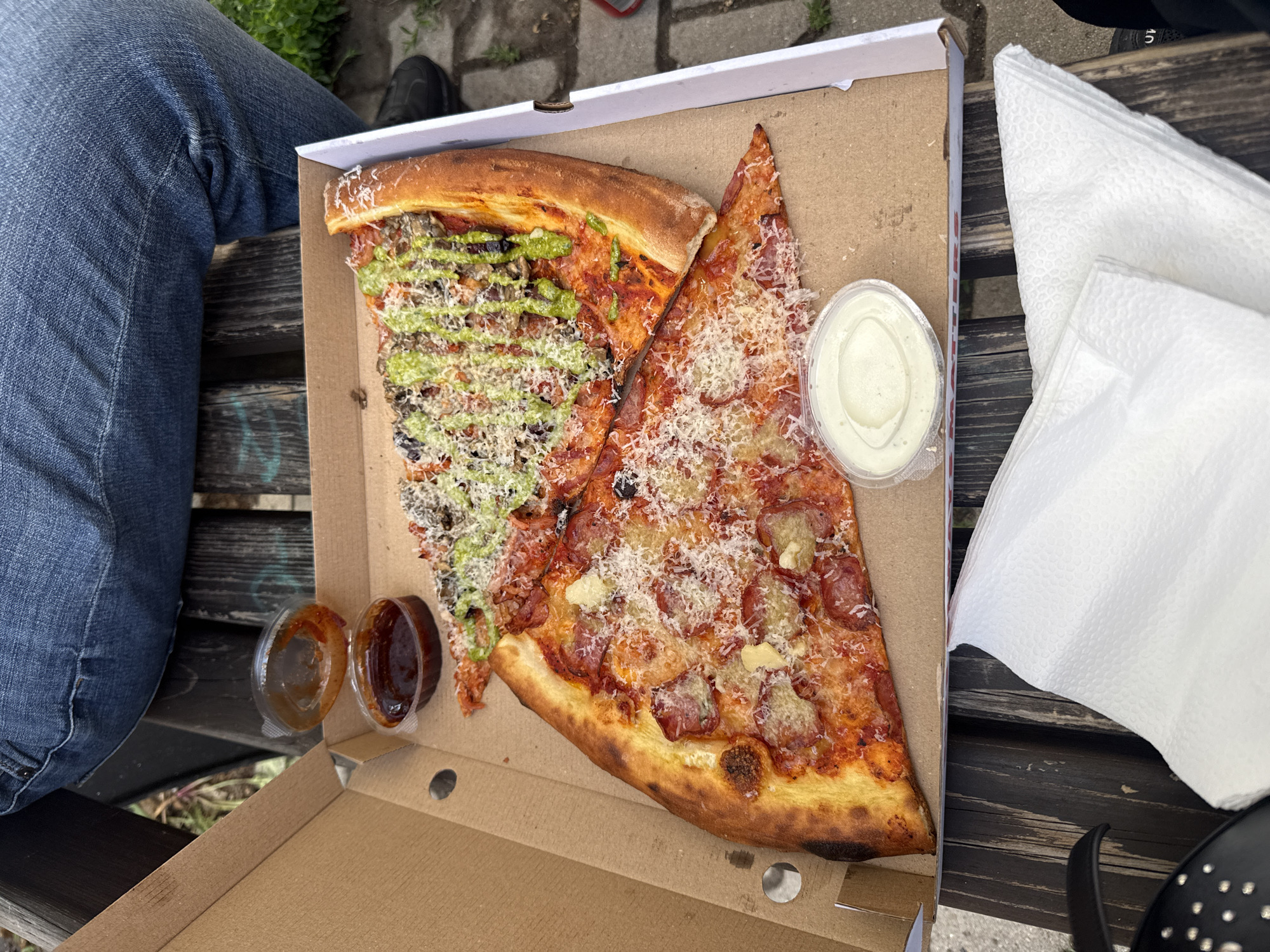 A partially-open pizza box rests on a dark-stained wooden bench, containing two slices of pizza, two small containers of brown sauce, and a small container of white sauce. One pizza slice is topped with green sauce, black olives, and cheese. The other is topped with pepperoni and cheese. A stack of white napkins sits to the right of the box. A person wearing blue jeans sits to the left. The bench appears to be outdoors, with concrete and grass visible near the legs of the person. The location is Sofia, Bulgaria.
