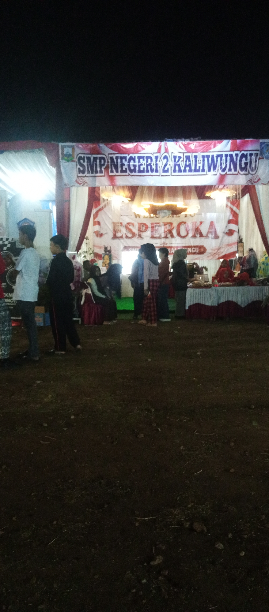 The image depicts an outdoor event at night, likely a school function, with a banner prominently displayed that reads "SMP NEGERI 2 KALIWUNGU" and "ESPEROKA". Several people are present, including children and adults, some standing and some seated. The background features a decorated stage area with draping and lighting. In the foreground, a patch of dark, uneven ground covered with some vegetation and scattered debris is visible. The overall atmosphere suggests a festive gathering. The presence of "SMP NEGERI 2 KALIWUNGU" indicates the event is associated with a public junior high school located in the town of Kaliwungu, Central Java, Indonesia.