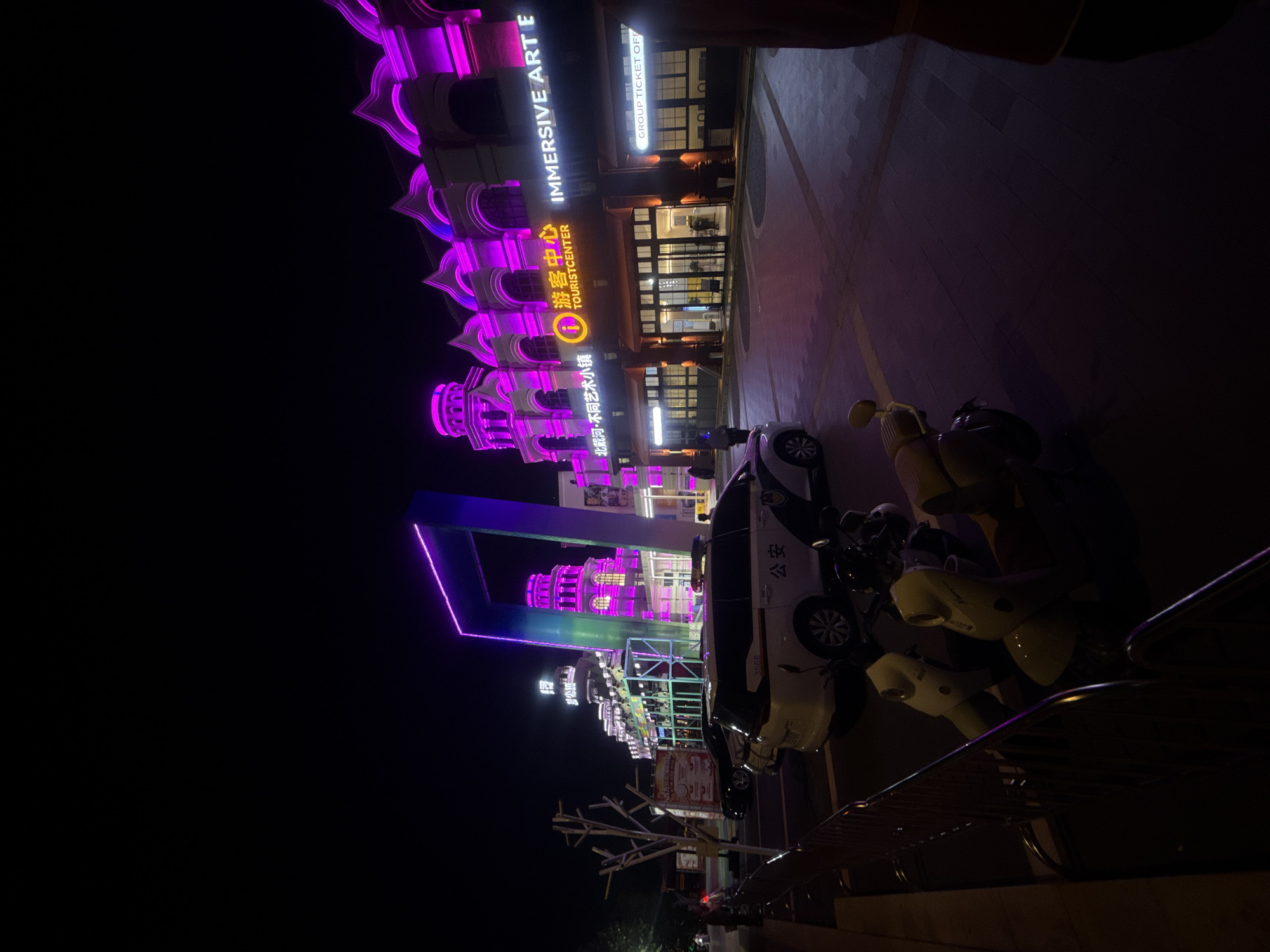 The image depicts a nighttime scene outside an establishment in Qinhuangdao, China, identified by signage. The building's facade is illuminated with vibrant pink neon lights, creating a striking contrast against the dark sky. In the foreground, several parked electric scooters are visible, with two prominent in the center of the frame. A police vehicle, marked with "公安" (Gōng'ān, meaning Public Security), is parked to the left of the scooters. The ground is paved with interlocking stones. The overall atmosphere suggests a commercial or entertainment area at night.