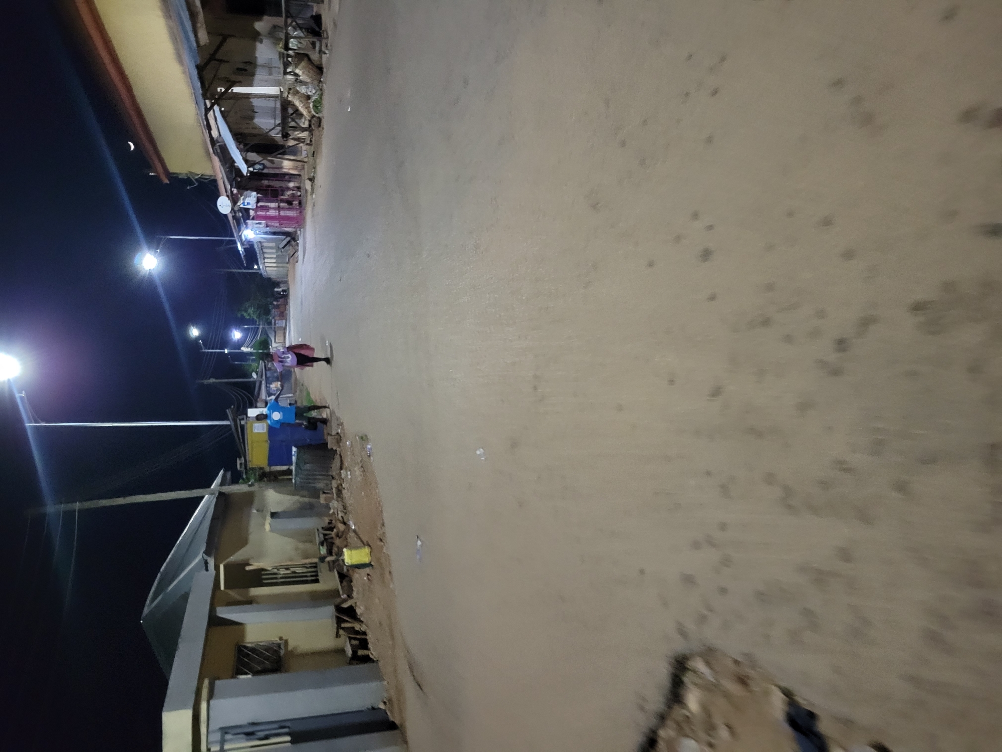 At night in an unknown city, the scene captures a long, slightly winding street illuminated by spaced-out streetlights. A person, blurred due to movement, is walking along the street. Buildings line the side of the road, some appearing to be shops or residences. The street surface itself is light-colored with scattered dark spots, possibly debris or stains. A crescent moon is visible in the dark sky. The overall setting suggests a quiet, possibly residential area.