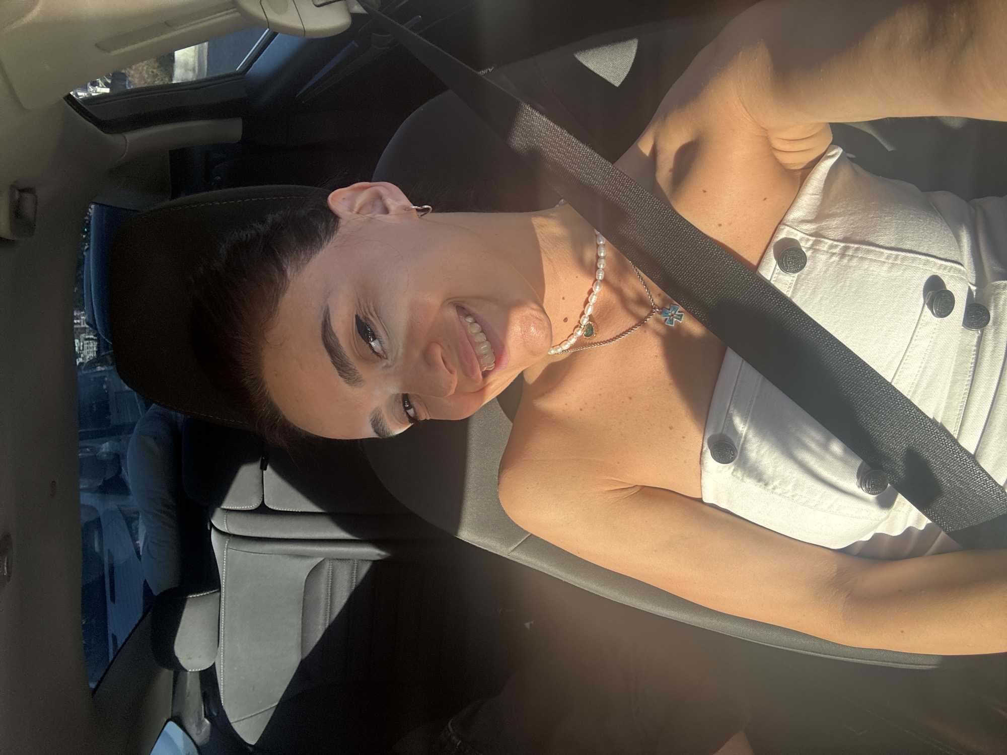 The image shows a woman inside a car, with the camera positioned at an upward angle. She is wearing a white strapless top and a pearl necklace with a blue cross pendant. A black seatbelt crosses over her chest. The woman has dark hair and is smiling, with her head tilted to the side. The interior of the car is visible, including a sunroof, black leather seats, and a part of the dashboard. Sunlight is streaming into the car, casting shadows on the seats and highlighting the woman's arm and face. The background outside the car window is blurred, but suggests an urban environment with buildings and trees. The lighting and shadows indicate it is daytime, likely a sunny afternoon. The setting appears to be Tbilisi, Georgia.
