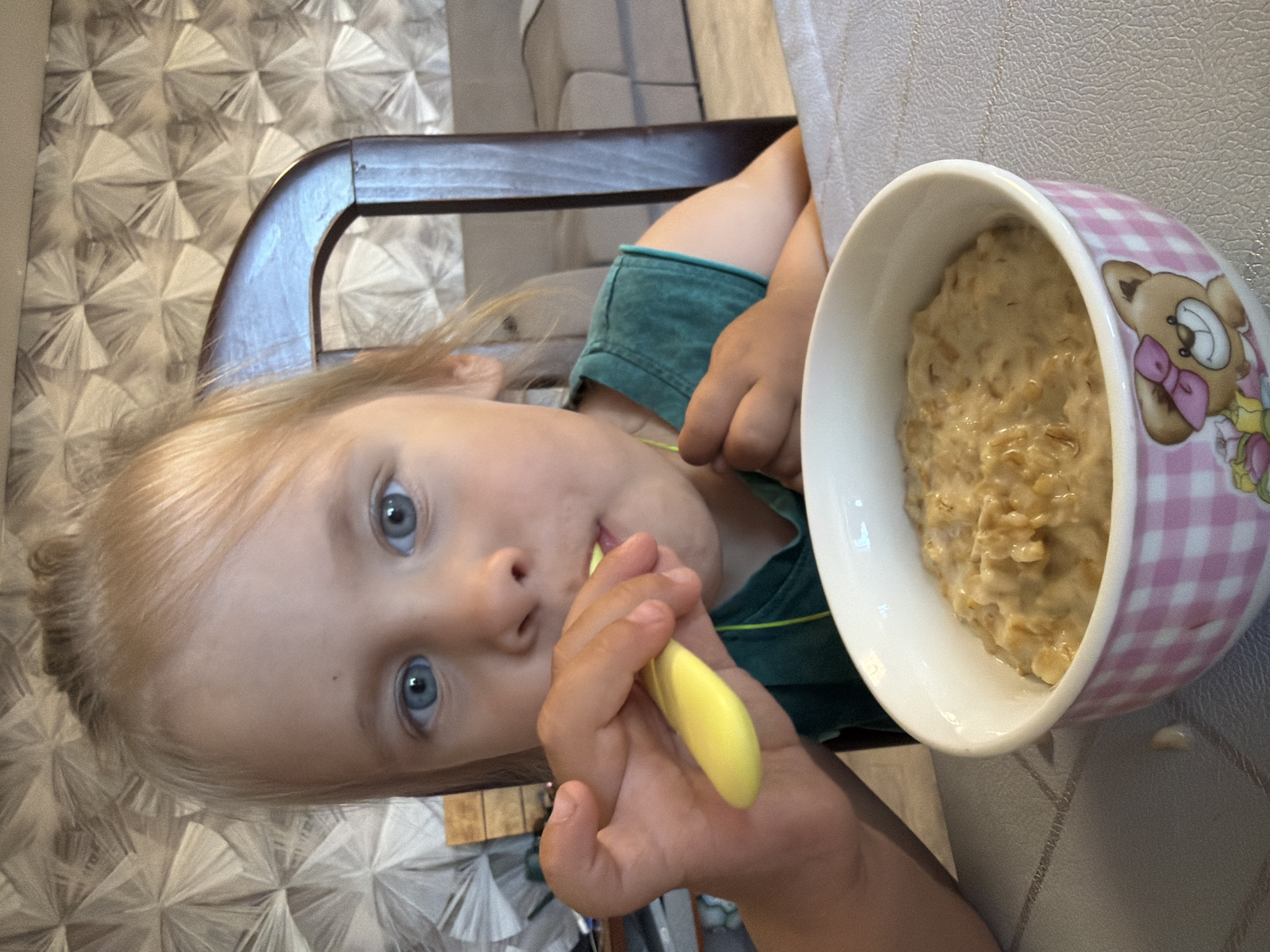 A close-up, eye-level shot shows a young child with blonde hair and blue eyes eating from a bowl of oatmeal. The child is seated at a table, holding a yellow spoon to their mouth. The bowl is pink and white checkered with a teddy bear design. In the background, a wooden chair and a textured wall are visible. The scene appears to be indoors, possibly in a home, in the city of Chaikovsky, Russia.