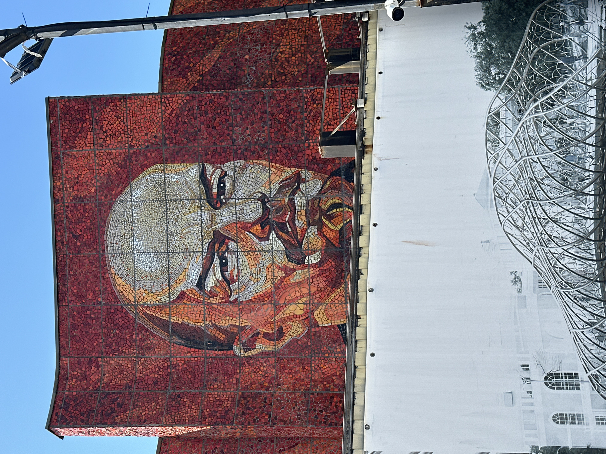 This media file presents a striking outdoor scene, dominated by a large, intricate mosaic adorning the facade of a building, likely situated in Sochi, Russia. The mosaic vividly depicts a stylized human face, possibly male, rendered in a dynamic palette of white, cream, orange, and brown tesserae, set against a rich background of red and dark red mosaic tiles. The face features expressive eyes and an open mouth, conveying a sense of intensity or contemplation. The individual tiles are irregularly shaped, contributing to the mosaic's textural depth and artistic quality.
Adjacent to the mosaic, a large white panel is affixed to the building. This panel features a black and white print of a contemporary architectural structure: an elaborate metallic lattice, possibly part of a dome or a decorative canopy, is visible in the foreground. Behind this modern element, a more traditional building with arched windows and a visible tree adds a contrasting layer to the printed image.
Above, a dark metal beam extends across the top of the frame, supporting various cables, zip ties, and a security camera, indicative of an urban environment. The bright, clear blue sky suggests a sunny daytime setting. The image primarily captures a static view of public art and architectural features, with no apparent activity taking place. No discernible text is visible within the frame.