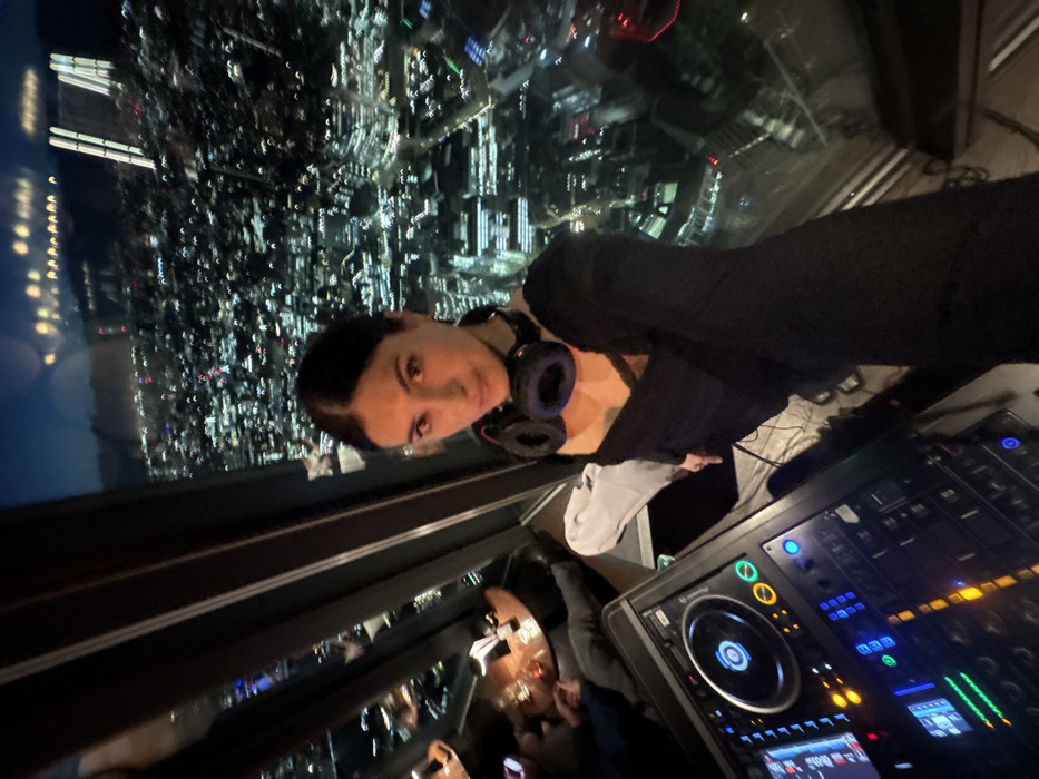 An elevated interior night scene from a high-rise building. The main subject, a female with dark hair pulled back, wearing a dark top and blue and black headphones around her neck, is positioned in the foreground, facing the camera. To her left, a professional DJ mixer/controller is partially visible, featuring illuminated buttons, faders, and a jog wheel. In the left background, behind the DJ equipment, indistinct figures are seated at small, round tables in a dimly lit lounge area. The expansive background reveals a blurred, illuminated cityscape at night, seen through large, slanted window panes. The structure of the windows and the high vantage point over the city of London indicate the location as a high-floor establishment within The Shard, London, United Kingdom.