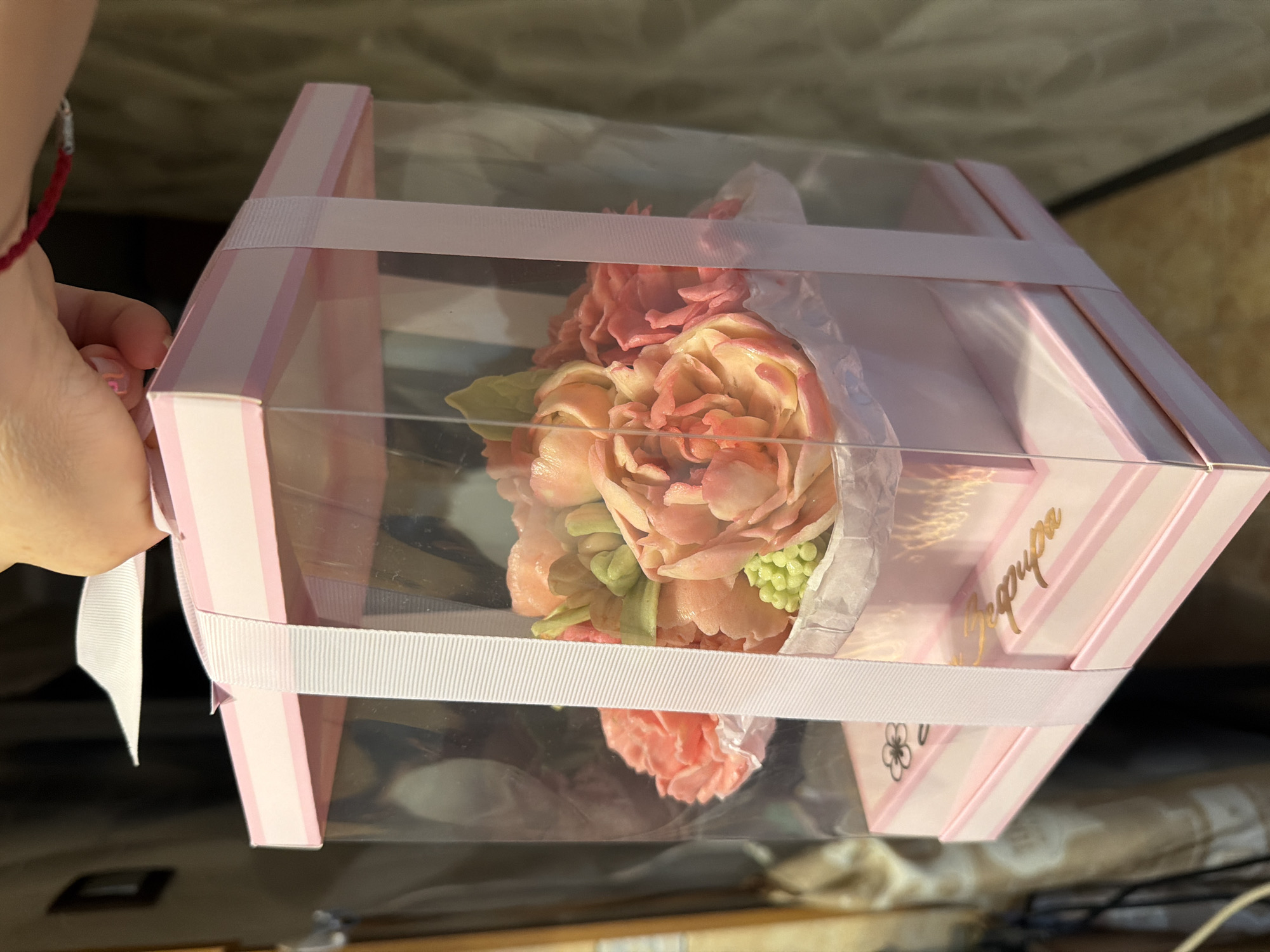 A hand holds a square-shaped, pink and white striped box with a clear plastic window, containing a bouquet of buttercream flowers. The box is secured with a wide, white ribbon wrapped around it. The text "Верфира" is visible in gold lettering on the side of the box, along with a small floral design. The image is taken indoors under ambient lighting. The exact location in Moscow, Russia cannot be determined from this image.