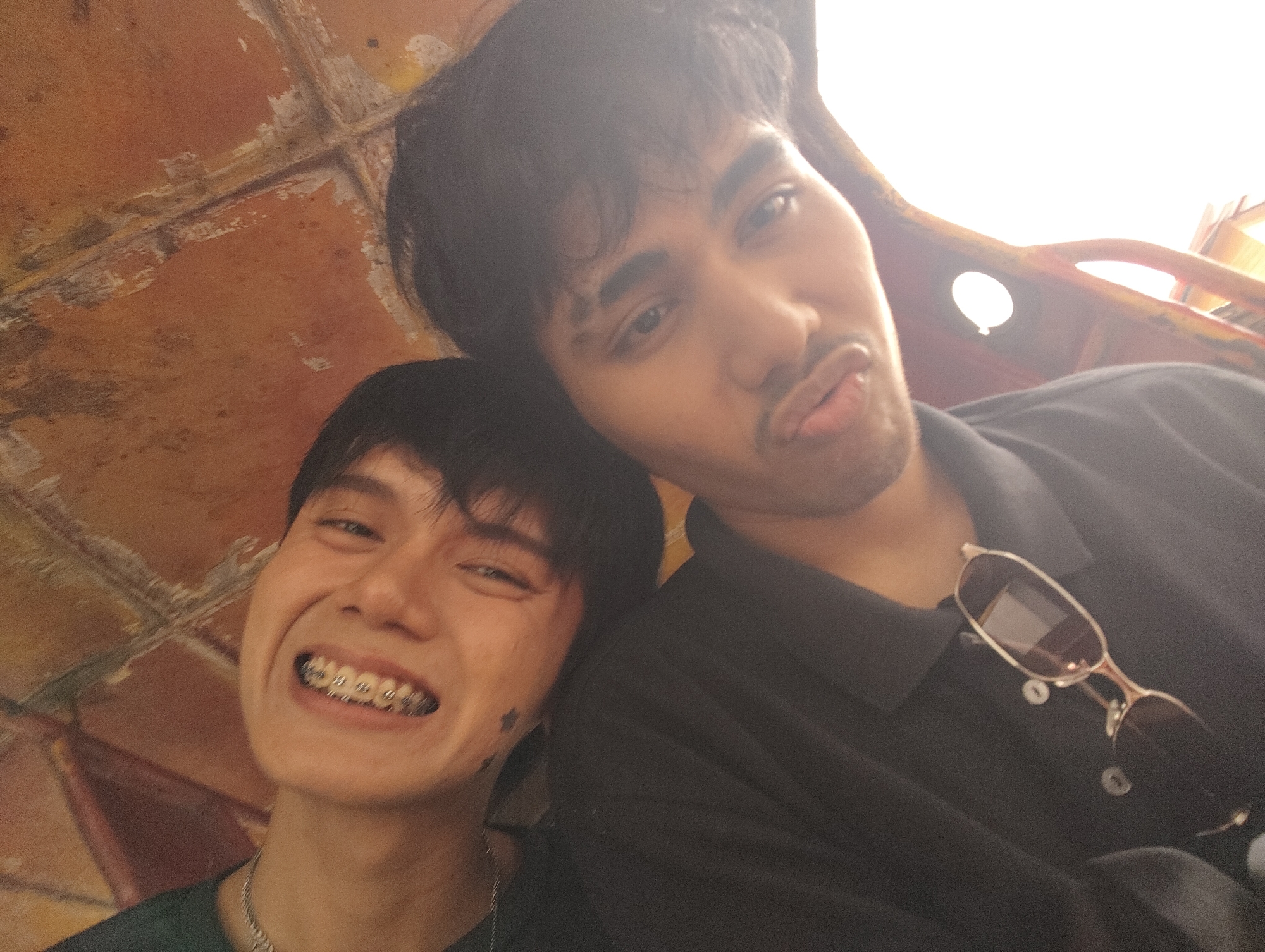 Two young men are taking a selfie, with the camera held by the person on the left. The man on the left has dark hair, is smiling broadly, showing off his braces, and is wearing a dark green shirt and a silver chain necklace. There is a small star tattoo visible on his left cheek. The man on the right has dark hair and is puckering his lips, giving a kissy face. He is wearing a black collared shirt and has sunglasses hanging from the collar. Both men appear to be in good spirits. The background suggests they are inside a structure with a textured, possibly aged, orange ceiling. The lighting is bright, with the right side of the image being overexposed, suggesting it may be daytime and they are near a light source or window. The overall atmosphere is casual and friendly.