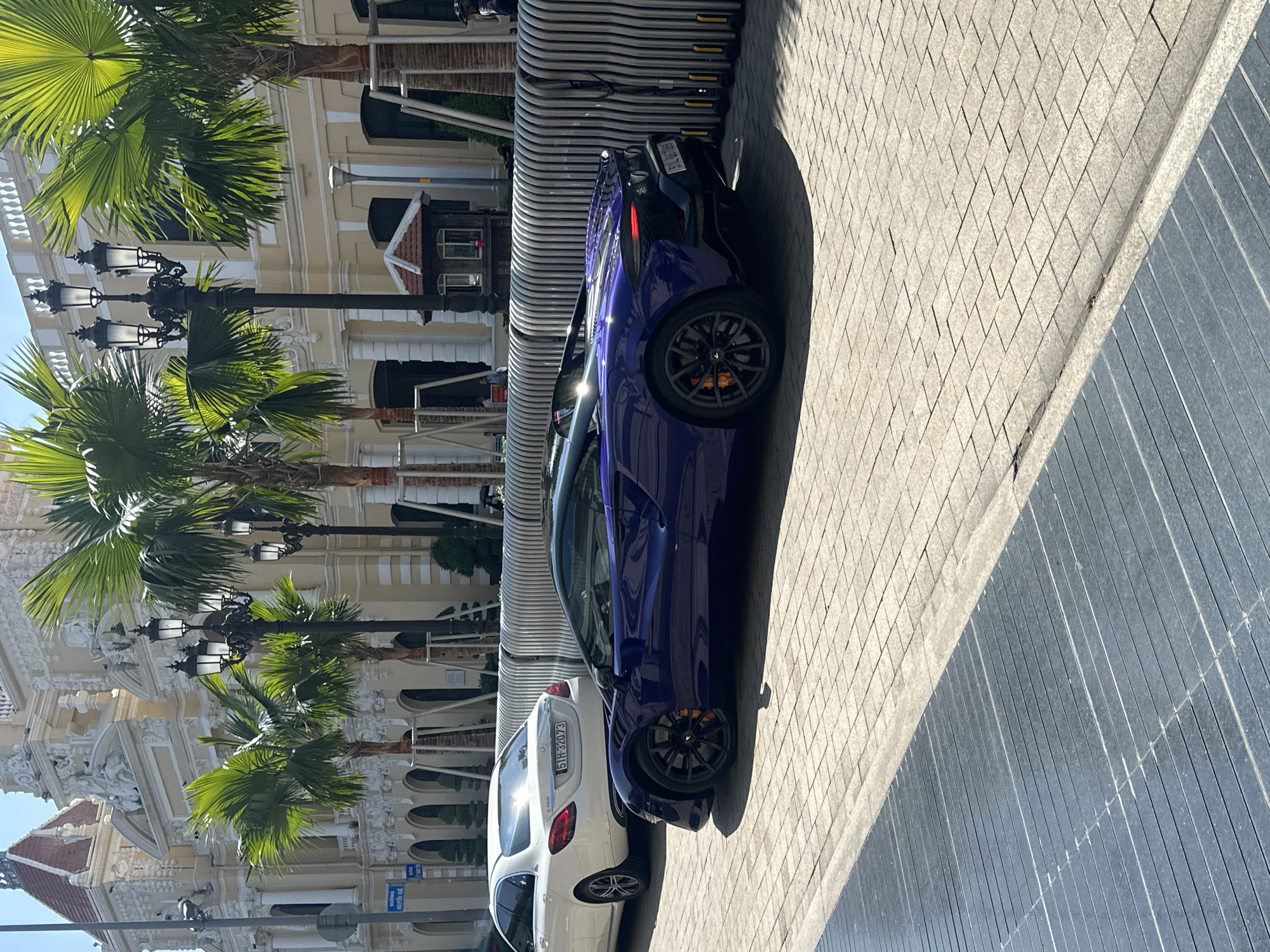 The image presents an outdoor scene in the city of Thủ Đức, Vietnam, featuring two vehicles parked along a brick-paved street. A white Mercedes-Benz C-Class with license plate "51H-320.73" is closely followed by a striking purple McLaren. The background reveals a classical architectural facade with tall, ornate columns, palm trees, and vintage-style street lamps. The scene suggests a blend of modern luxury automobiles juxtaposed with a historic urban environment.