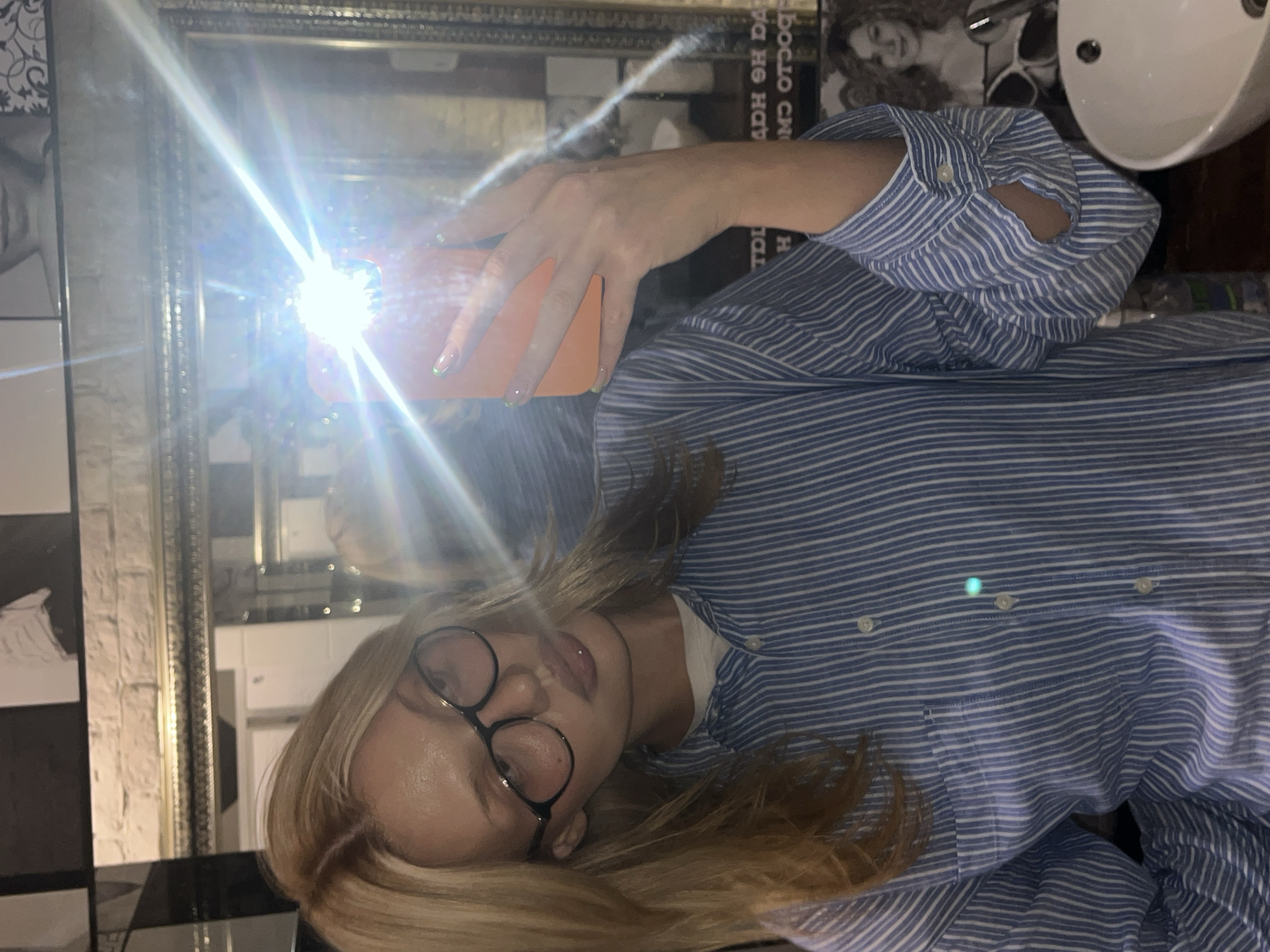 A blonde-haired woman wearing round glasses and a blue and white striped shirt takes a selfie in a bathroom. She is holding an orange phone, with the flash obscuring the background details behind the mirror. A white sink is visible in the upper right corner. The woman appears to be in a bathroom with black and white tiled walls. The location is Chaikovsky, Russia.