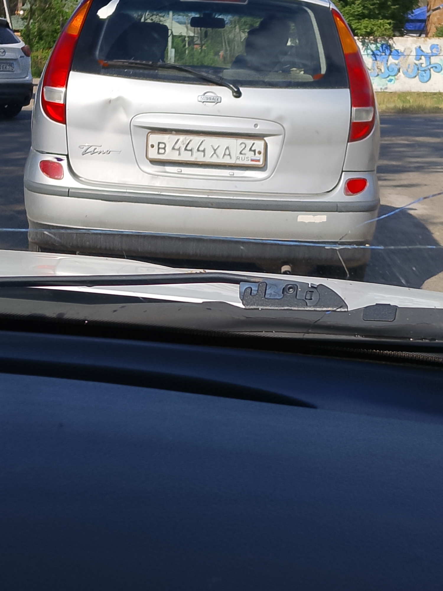 The media file shows the rear of a silver Nissan Almera Tino, likely from the early 2000s, parked outdoors on a sunny day. The car has sustained damage to its rear, with a noticeable dent in the center of the tailgate. The license plate is clearly visible and reads "B 444 XA 24 RUS". To the left of the license plate, the "Tino" model name is embossed on the car's body. The car is parked on a paved surface, with a damaged white wall covered in graffiti visible in the background to the right. Green foliage is present behind the wall, suggesting a semi-urban or suburban setting. The camera appears to be positioned inside another vehicle, looking out through the windshield, with parts of the interior, including the dashboard and windshield frame, visible in the foreground. The sunlight suggests it is daytime, and the clear visibility indicates fair weather. The overall scene depicts a car that has been in an accident or collision.