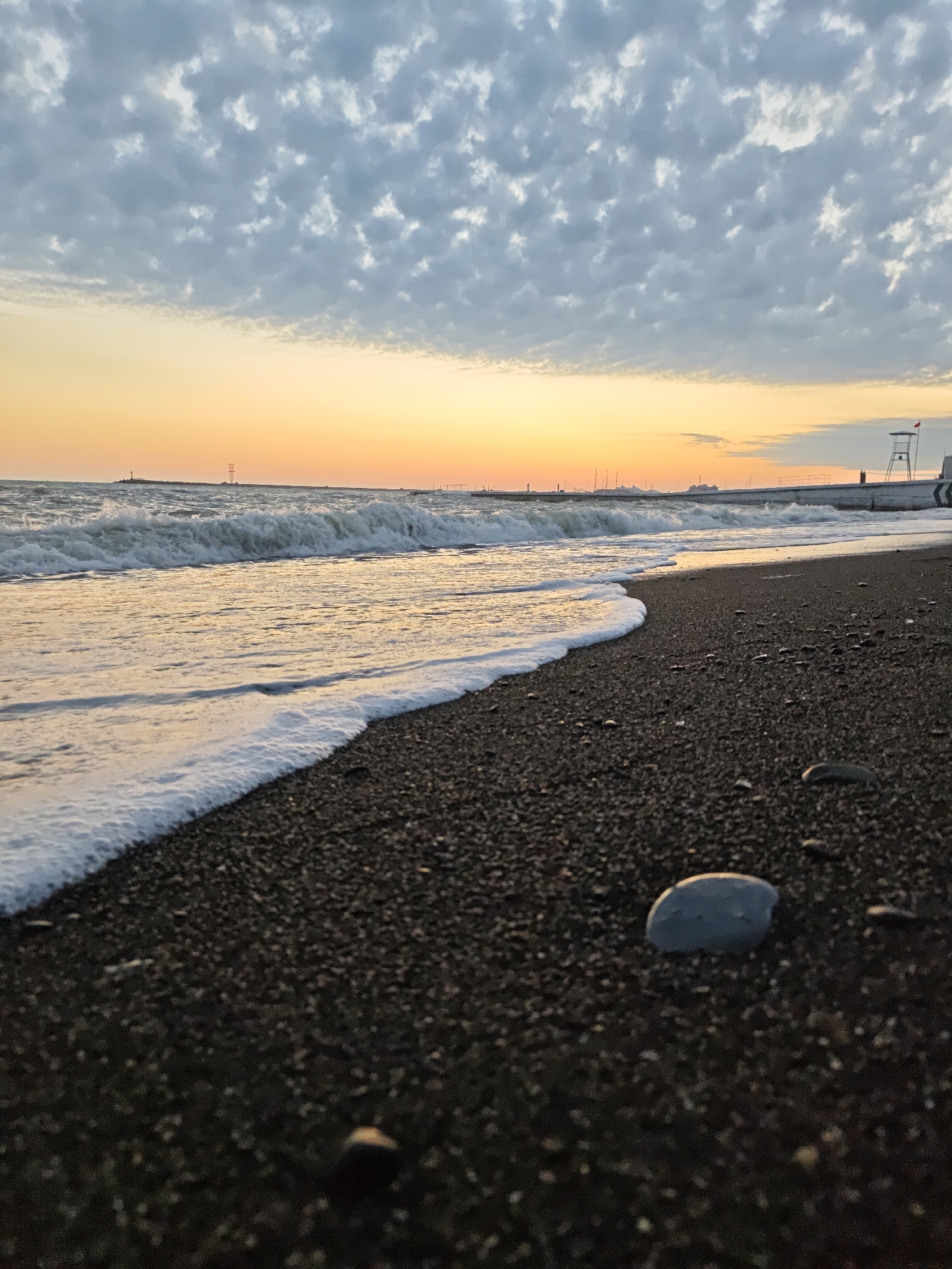 This image depicts a beach scene in Sochi, Russia. The foreground features a dark, pebble-covered beach with a small, light-colored stone prominently displayed. The shoreline is marked by gentle waves edged with white foam. In the midground, the sea extends towards the horizon, where a yellow-orange sunset illuminates the sky, partly obscured by a layer of white clouds. In the distance, there is a breakwater with various structures, including a lookout tower and utility poles. The overall scene captures a serene and coastal environment at dusk.
