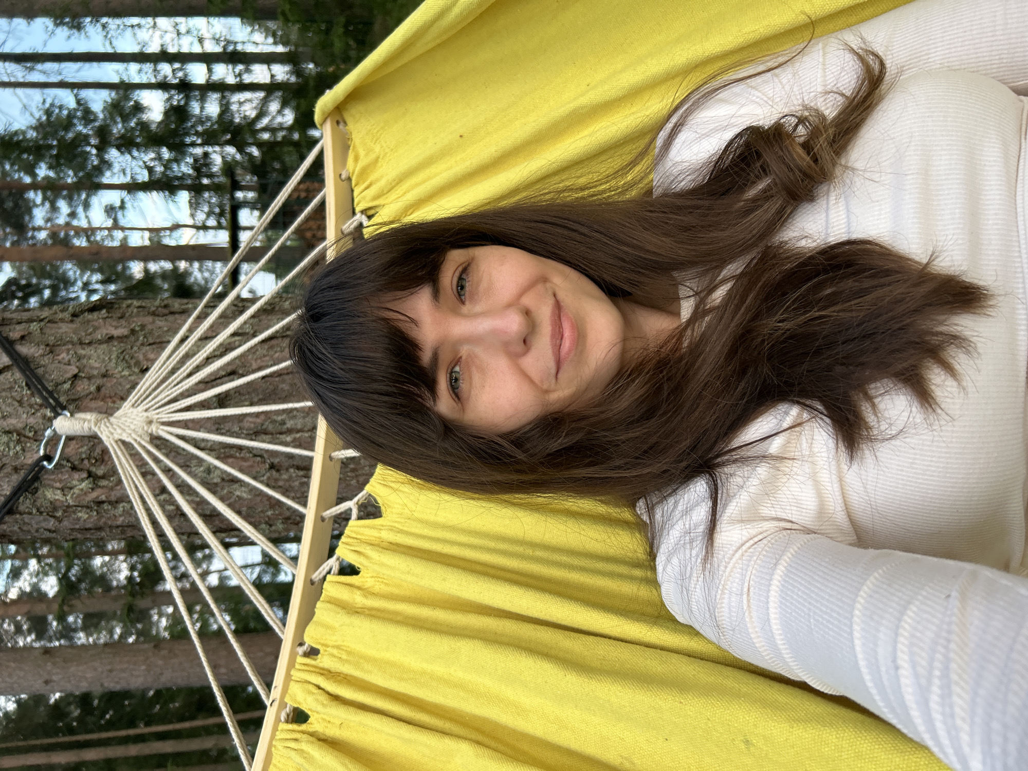A medium shot shows a woman with long, dark brown hair and bangs, looking directly at the camera with a slight smile. She is lying in a yellow hammock, which is suspended by ropes attached to a tree trunk. The woman is wearing a white, long-sleeved, ribbed top. The background is a forest with tall, slender trees and a blue sky visible through the branches. The location is Shepelyovo, Russia.