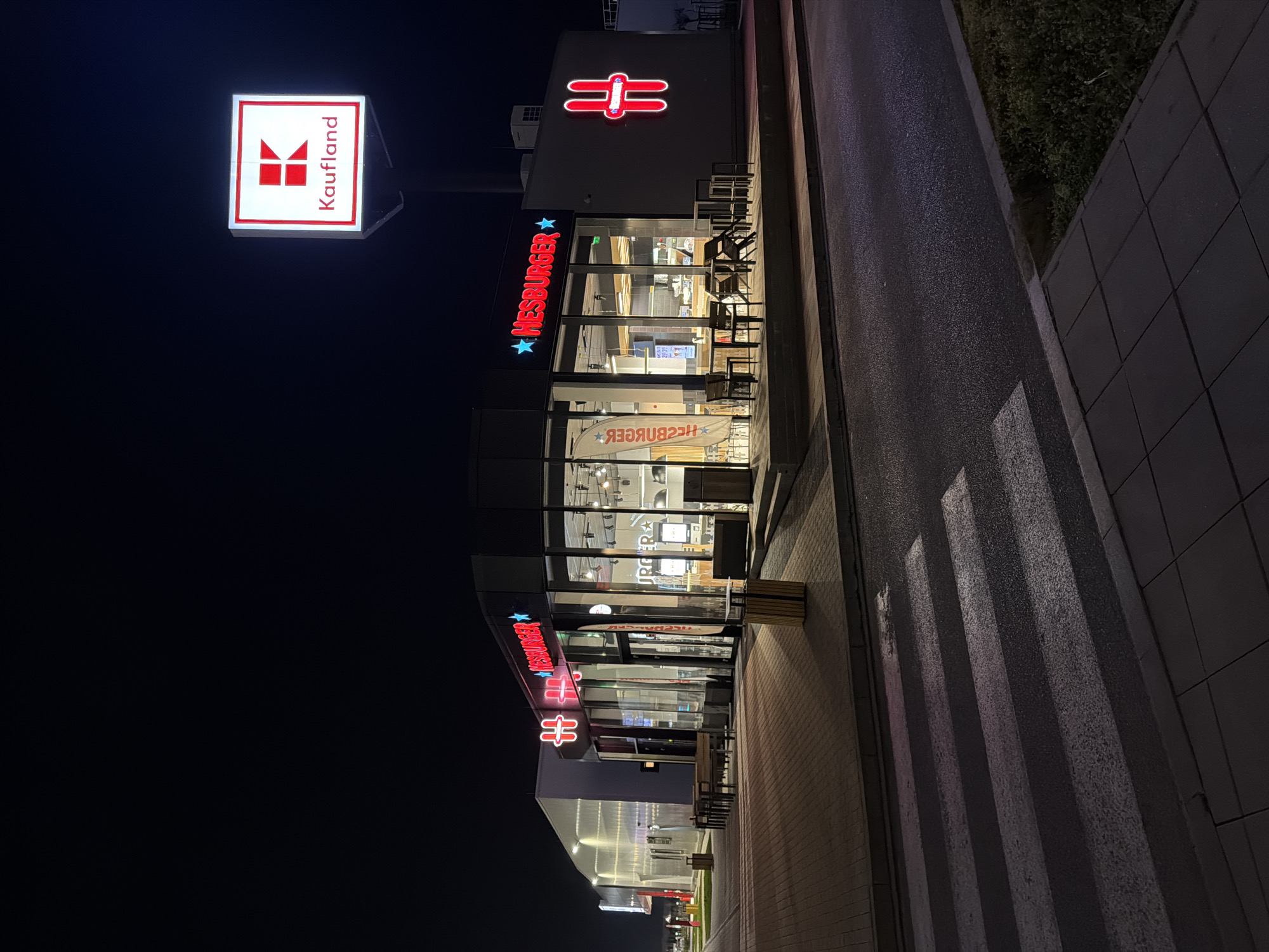 The image shows a Kaufland hypermarket and a Hesburger restaurant at night. The Kaufland logo is visible on a sign, and the Hesburger logo is displayed on the restaurant building. The scene is dark, with the illuminated signs and building providing the primary light sources. There are crosswalk stripes on the pavement near the restaurant. Based on the presence of both Kaufland and Hesburger, this image likely originates in a European city. Without the explicit knowledge of the location in Sofia, Bulgaria, it's impossible to know if the image is indeed taken in this particular city.