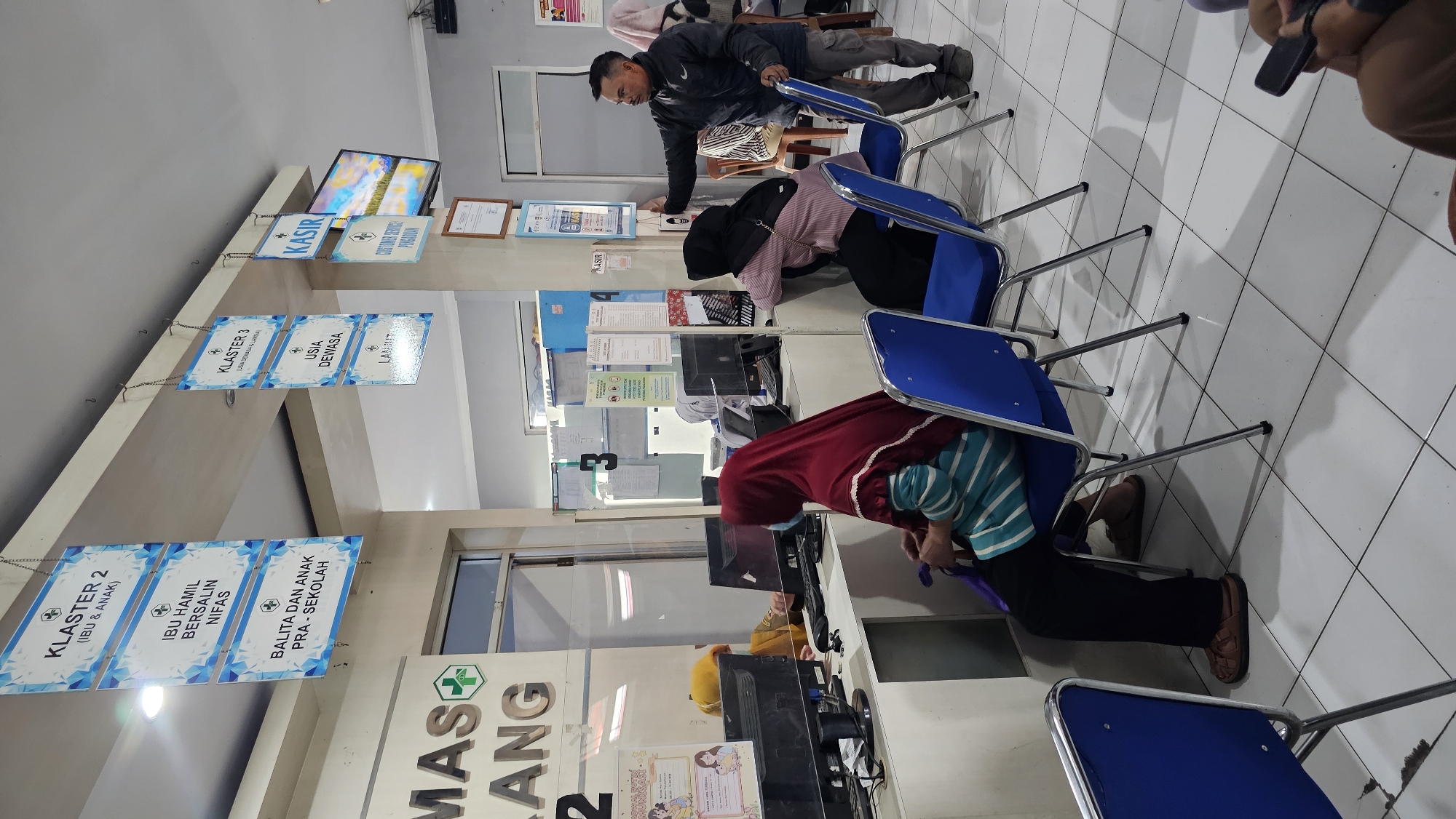 The image depicts an indoor waiting area, likely a clinic or government office, with several people seated and standing. In the foreground, a woman wearing a red hijab and a striped shirt sits on a blue chair, facing away from the camera. To her right, another woman in a pink and black striped top and black hijab is also seated, facing the counter. A man in a dark jacket and grey pants stands near the counter, appearing to interact with someone.
Several signs are hung from the ceiling. One prominent sign reads "KLASTER 2 (IBU & ANAK)" followed by "IBU HAMIL BERSALIN NIFAS" and "BALITA DAN ANAK PRA-SEKOLAH". Another set of signs includes "KLASTER 3 (USIA DEWASA & LANIA)", "USIA DEWASA", and "LANJUT". A sign for "KASIR" (cashier) is also visible, with a television screen displaying a landscape scene behind it.
The environment features a tiled floor, a long counter with multiple computer monitors, and fluorescent lighting. The overall impression is one of an organized public service area. The presence of the signs suggests that this location is in Kertamulya, Indonesia, as indicated by the context.