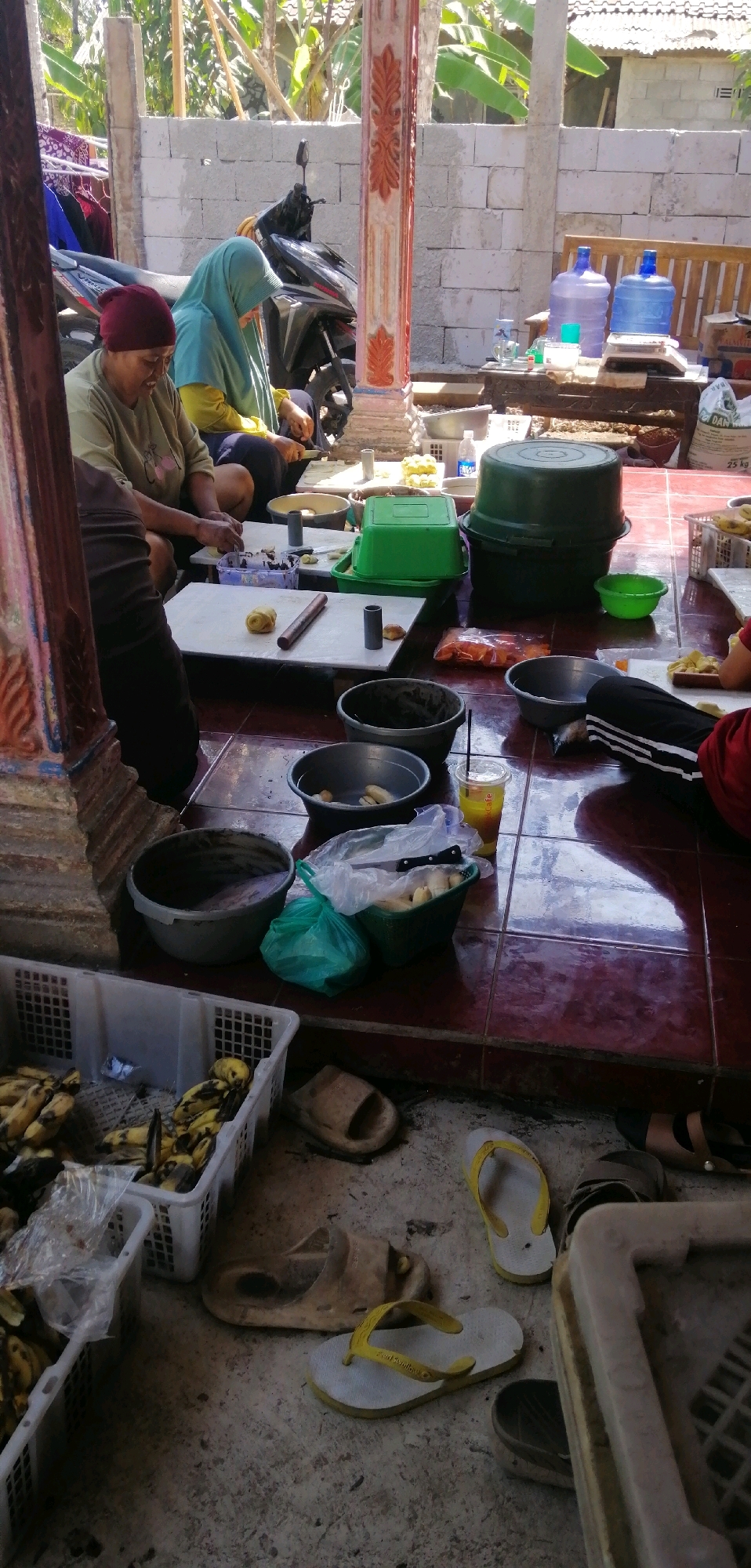 In a semi-open outdoor area with a tiled floor and cinder block walls, two individuals are engaged in food preparation. To the left, a person wearing a maroon head covering and a tan shirt is seated and working with dough on a white mat, with a rolling pin nearby. Beside them, another individual in a turquoise hijab and yellow top is also seated and appears to be processing dough. Several containers, including green plastic bins, large dark green tubs, and various bowls, are scattered around the workspace. In the foreground, white plastic baskets filled with bananas are visible, along with several pairs of sandals on the floor. A motorcycle is parked in the background, and sunlight illuminates the scene. The presence of water dispensers and a bag labeled "DAN" suggests this may be a commercial or community food production setting in Temanggal, Indonesia.