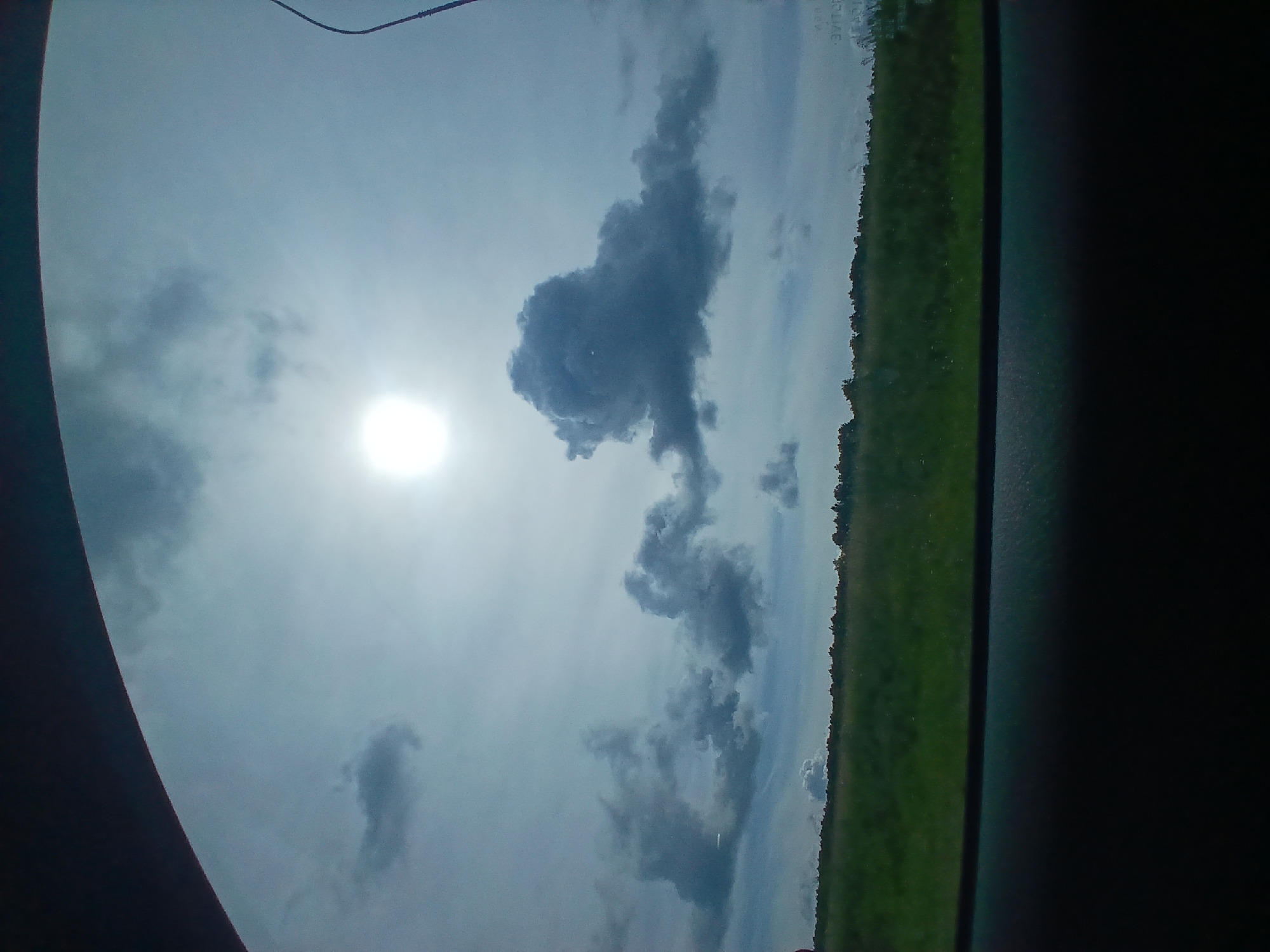 The image shows a view through a car window of a landscape under a cloudy sky with the sun partially visible and shining brightly. The foreground is dark, indicating the interior of the car. The middle ground features a grassy field with a line of trees in the distance. The sky occupies the upper portion of the image and is filled with various cloud formations, with the sun appearing as a bright, slightly obscured orb. The overall impression is a scenic view from a moving vehicle, likely on a highway or country road. There are no people or specific structures visible that would indicate a particular location within a city.