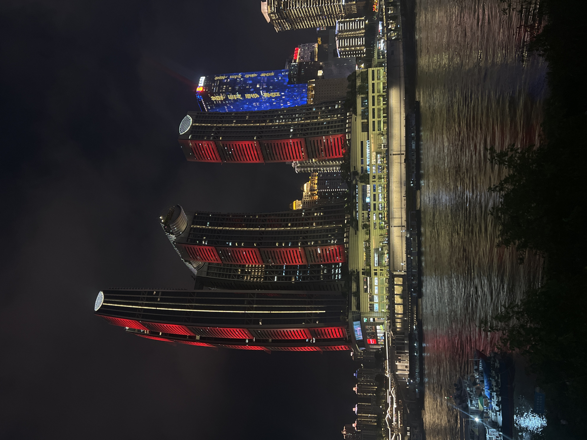 This image captures a night scene of the Jiangbei Cheng Street in China, featuring a striking modern cityscape. Dominating the view are several illuminated skyscrapers, with the most prominent one on the left showcasing a dynamic facade of red and white vertical lighting. Adjacent to it, another tall building displays a vibrant blue light show, with golden characters visible on its surface. The buildings are situated along a wide river, which reflects the colorful lights of the city. In the foreground, a section of the river is occupied by several boats, some of which are docked and illuminated. The riverbank is lined with a multi-story building complex, also lit up, suggesting a commercial or entertainment area. The sky above is dark, with hints of clouds, emphasizing the artificial illumination of the urban environment. The overall impression is one of a bustling, modern metropolis at night.