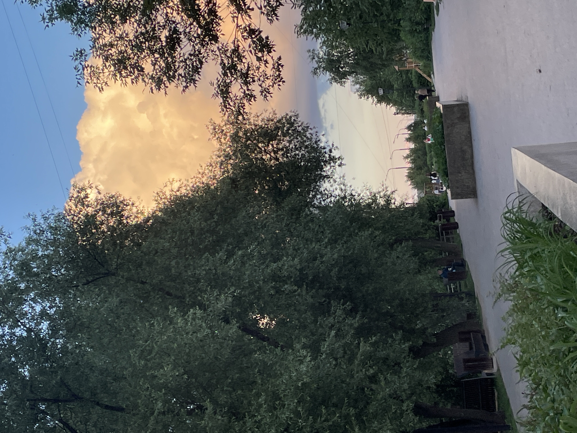 The image depicts a park scene in the city of Perm, Russia, likely during the late afternoon or early evening, as indicated by the warm, golden light illuminating a large cloud in the sky. The foreground features a paved walkway lined with lush green foliage and decorative concrete planters. To the left, a row of mature trees with dense, dark green foliage casts shadows onto the walkway. Beyond the trees, several park benches are visible, with one person seated on a bench. Further down the walkway, a group of people are scattered throughout the park, some walking, some standing. A playground structure is visible to the right. The sky transitions from a pale blue at the top to hues of pink and orange near the horizon, where a prominent, brightly lit cloud dominates the upper portion of the frame. Power lines are faintly visible across the sky. The overall atmosphere is serene and suggests a public space enjoyed by residents.