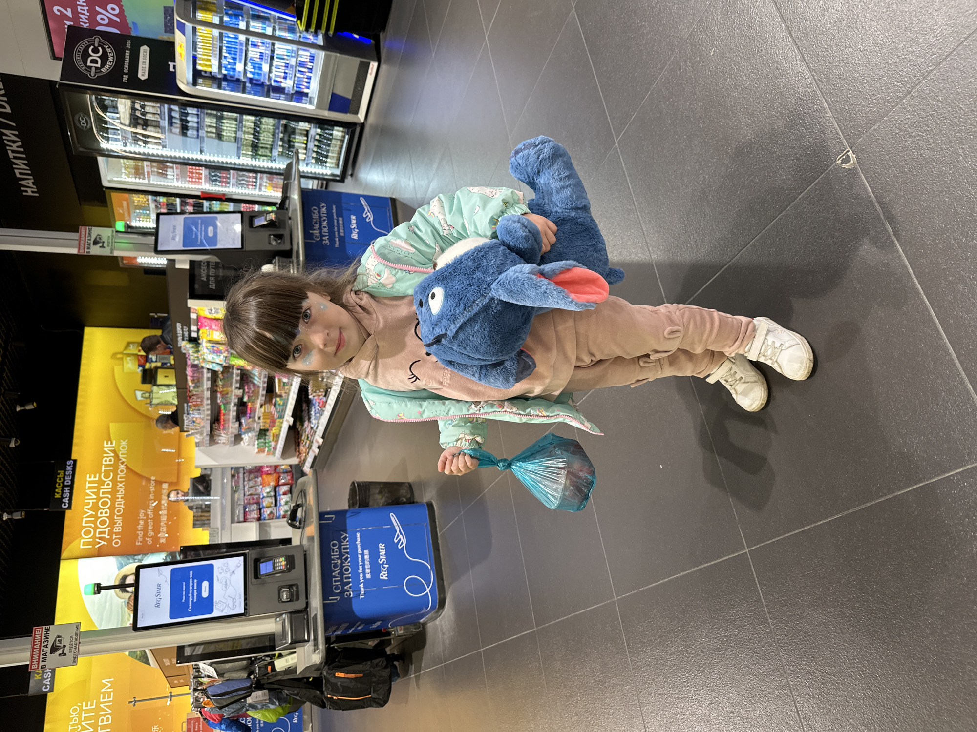 A young girl with glitter on her face stands in a grocery store in Sochi, Russia, holding a plush toy and a plastic bag. She wears a light pink outfit with a patterned jacket and white sneakers. The store has self-checkout kiosks and refrigerators filled with drinks. Advertisements in Russian and English are visible. The floor is covered with dark gray tiles.