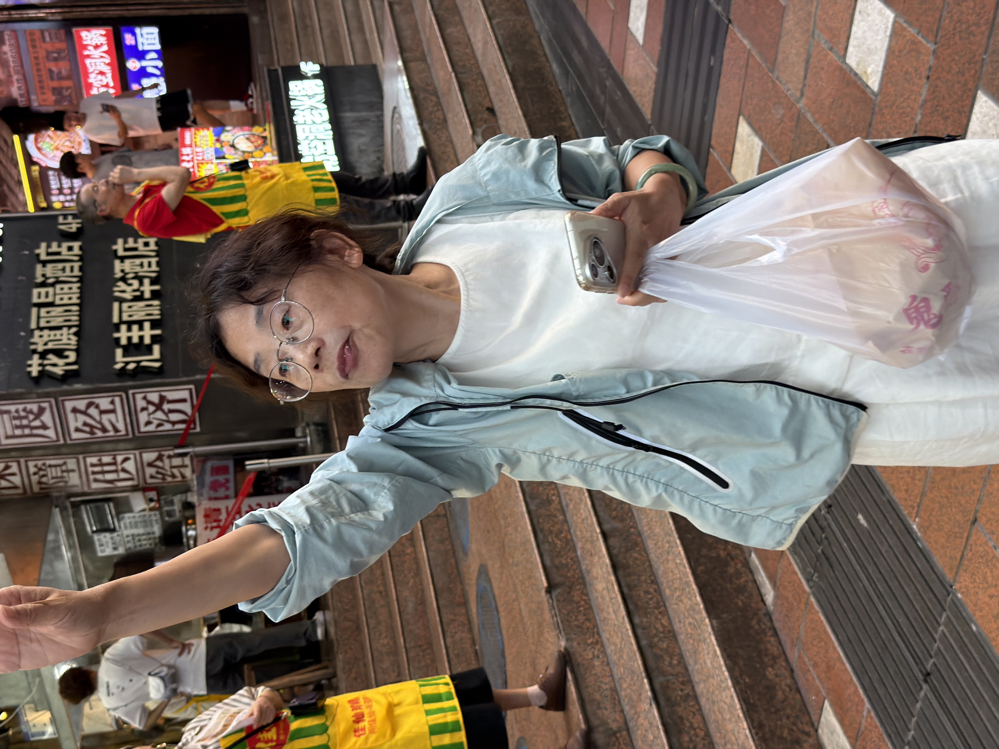 An Asian woman in her 50s stands on a sidewalk in Yuzhong District, China. She has dark, curly hair and wears glasses, a light blue jacket, and a white top. Her right arm is raised, and she holds a smartphone and a translucent plastic bag containing items. A green bracelet is visible on her left wrist. Behind her, a man in a red and yellow apron stands on a set of stairs, and other individuals are visible in the background. The area appears to be a commercial street with neon signs and shop fronts. The ground is paved with reddish-brown tiles and a metal grate.