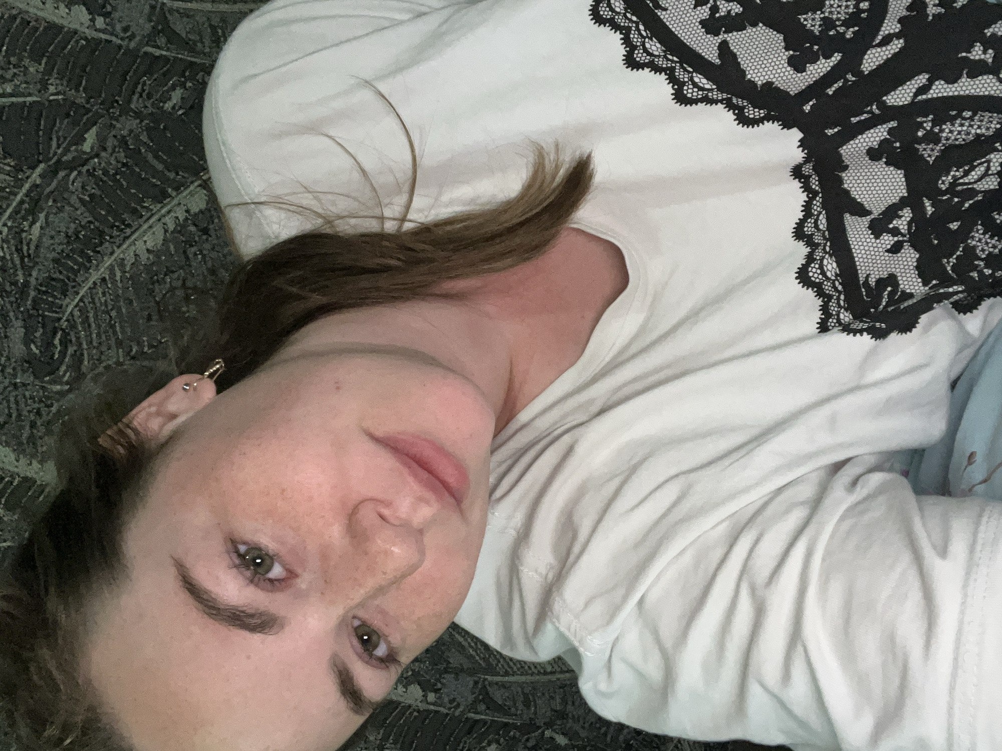 A woman lies on a dark, patterned surface, possibly a rug or bedspread, wearing a white t-shirt with a black lace-like design on the upper right. She has light brown hair and fair skin with freckles. Her eyes are light-colored. An earring is visible in her left ear. The background texture is nondescript, with no indication of being in the city of Chaikovsky, Russia. There is nothing indicating any specific actions or interactions.