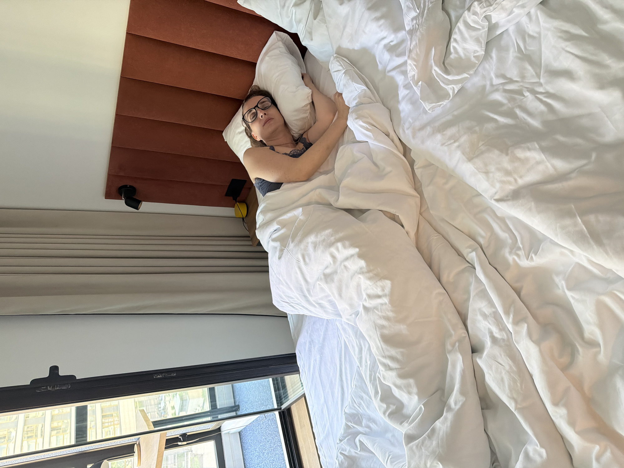 A woman wearing glasses and a blue top sleeps in a bed covered in white sheets and a white pillow. The bed has a brown headboard. A window is visible, revealing a view of buildings outside, and light fixtures are attached to the headboard. The location is Khimki, Russia.