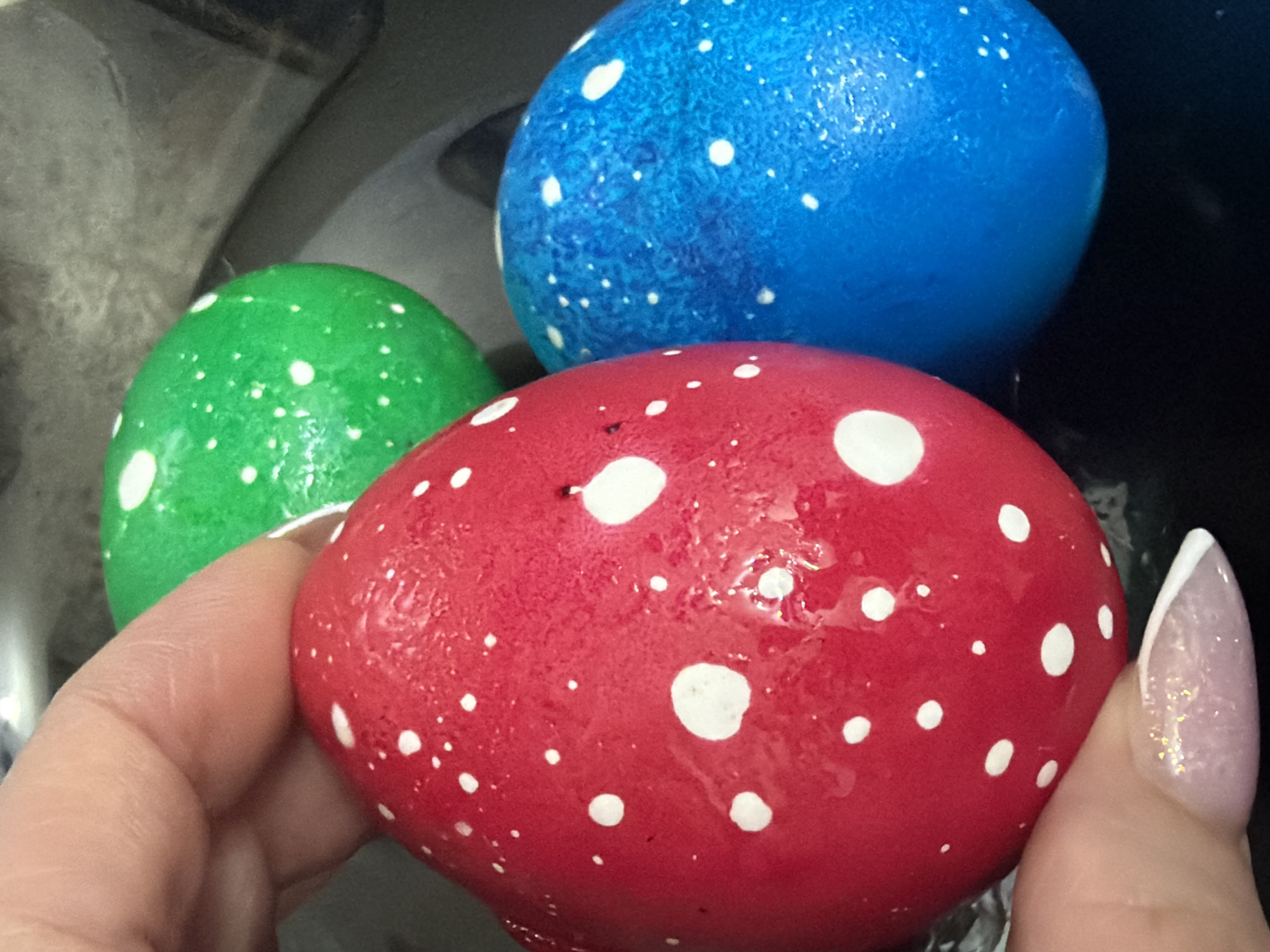 The image depicts three brightly colored Easter eggs. The eggs are dyed green, red, and blue, and each has white polka dots. A hand with an elongated manicure, painted light pink with white tips, holds the red egg. The background is indistinct. It is not possible to infer the location.