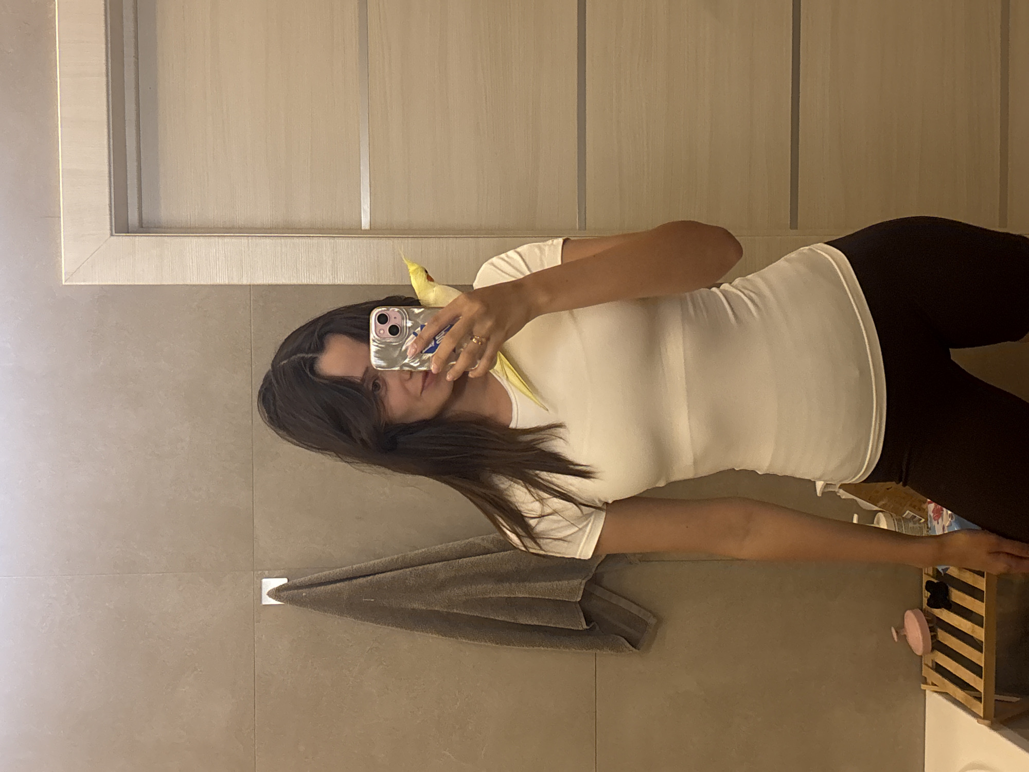 A young woman with dark hair and bangs takes a mirror selfie in what appears to be a bathroom. She is wearing a white t-shirt and dark leggings. A yellow cockatiel is perched on her shoulder, peeking out from behind her head. The bathroom features beige tiled walls, a towel hanging on a hook, a wooden shelf with bath accessories, and a light-colored door in the background. There is no specific information that would allow for the identification of Krasnoyarsk, Russia as the location.