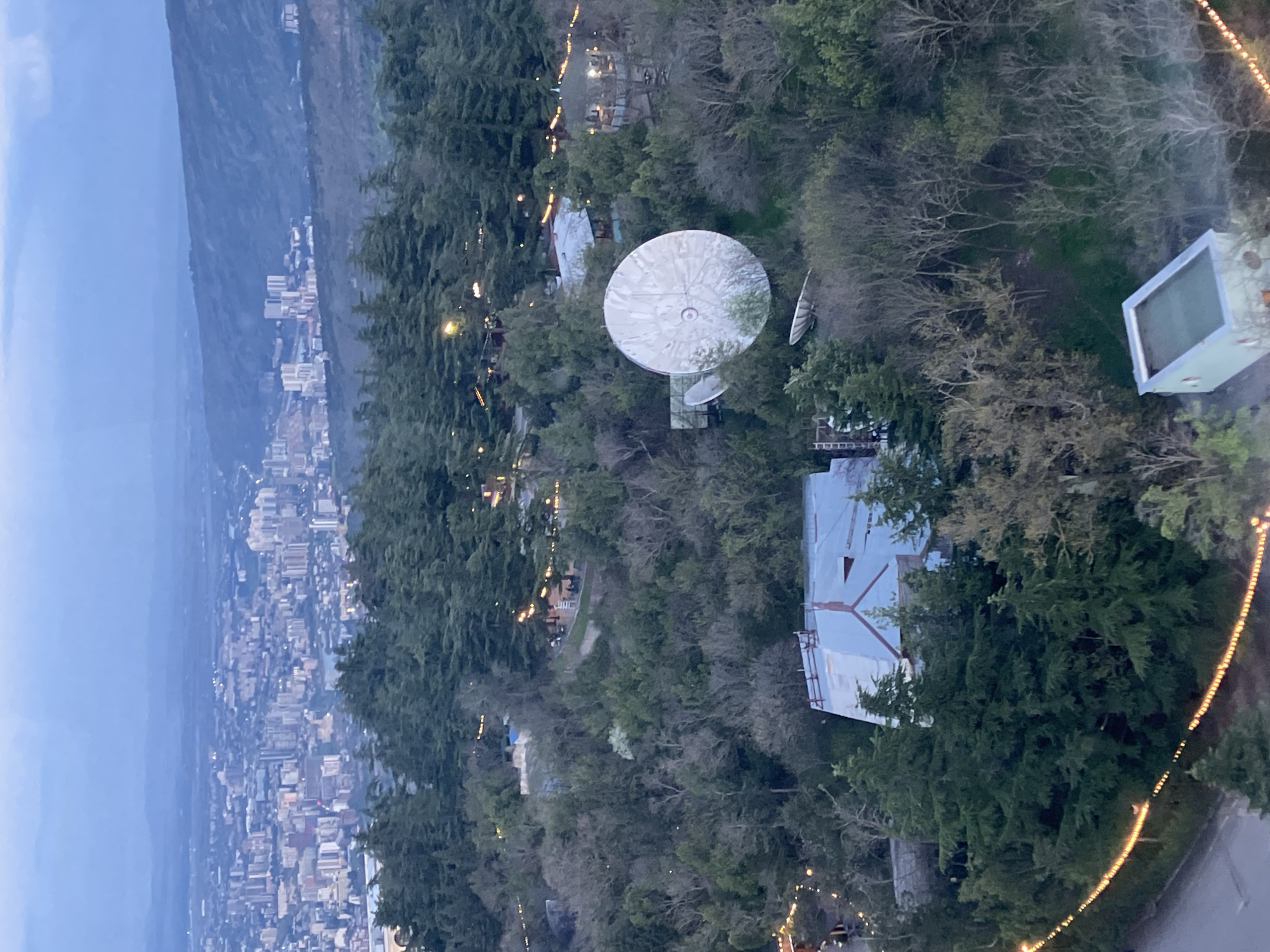 The image presents a high-angle, panoramic view of a landscape at dusk or early night, looking down over forested hills towards a sprawling city.
In the immediate foreground and mid-ground, dense green trees dominate, covering the hilly terrain. Among the trees, several structures are visible. A large, prominent white satellite dish, circular in shape, stands out, surrounded by foliage, with at least one smaller dish nearby. Other buildings with light-colored, possibly metallic or reflective, roofs are interspersed among the trees; one features a distinct gabled roofline. String lights with warm yellow bulbs are strung through parts of the forested area, suggesting paths, outdoor seating, or recreational spaces. A rectangular, light-colored structure, possibly a swimming pool or a large container, is also seen amidst the trees.
The background reveals a vast urban area, likely Tbilisi, Georgia, extending across a valley or along hills. Numerous buildings are packed together, with their lights beginning to illuminate the cityscape. Beyond the city, the silhouettes of distant mountains or hills merge with the deep blue-grey twilight sky. The overall impression is one of a quiet evening view, with artificial lights starting to define the human presence against the natural landscape. No discernible human activity or text is visible within the frame.