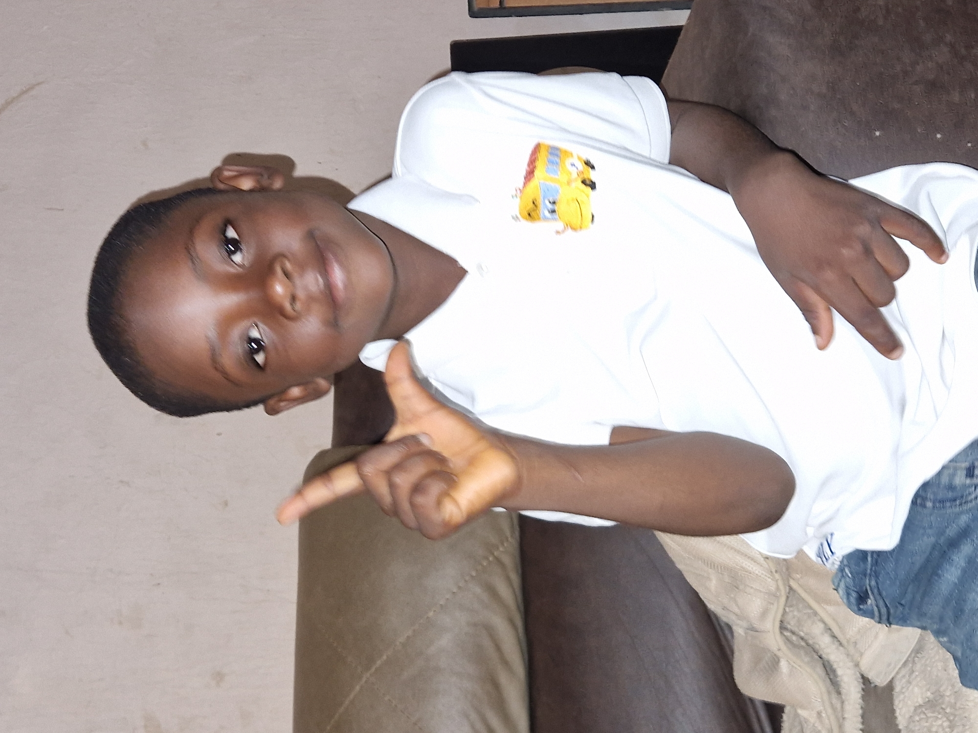 A young boy of African descent, with dark skin and short, dark hair, is pictured indoors. He is wearing a white polo shirt with a cartoon yellow school bus emblem on the left chest and blue jeans. The boy is seated on a dark brown sofa, with a lighter colored blanket or throw draped over the left side of the sofa. A light-colored wall serves as the backdrop. The boy is looking towards the camera with a slight smile and is making a hand gesture with his right hand, extending his index and middle fingers upwards, while his thumb touches his ring finger, forming an "L" shape. His left hand rests on his lap. The lighting appears to be artificial, casting a shadow on the wall behind him. The image quality is moderate, with some noise and a slight blur. The scene does not contain any specific identifiable landmarks or clues to pinpoint its exact location within Jos, Nigeria, beyond it being an indoor setting.