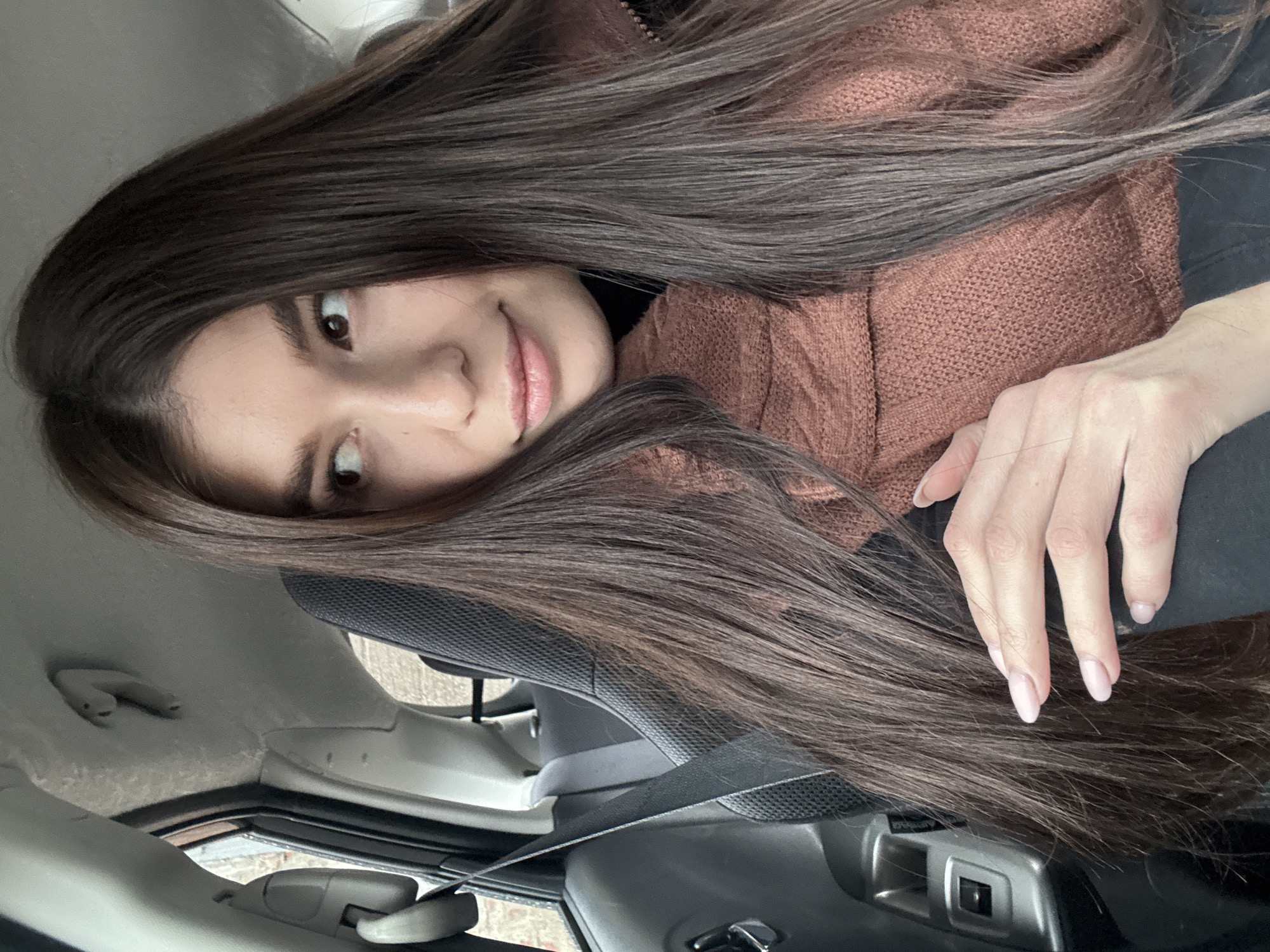 A young woman with long, dark brown hair is positioned inside a car. She is wearing a brown sweater, and her hair is partially obscuring her face and shoulder. Her right hand is visible, with light pink nail polish. The car's interior includes the seat, seatbelt, dashboard, and ceiling. The location is implied to be Chaikovsky, Russia, but there are no visual elements to explicitly confirm the city or country in the photo.