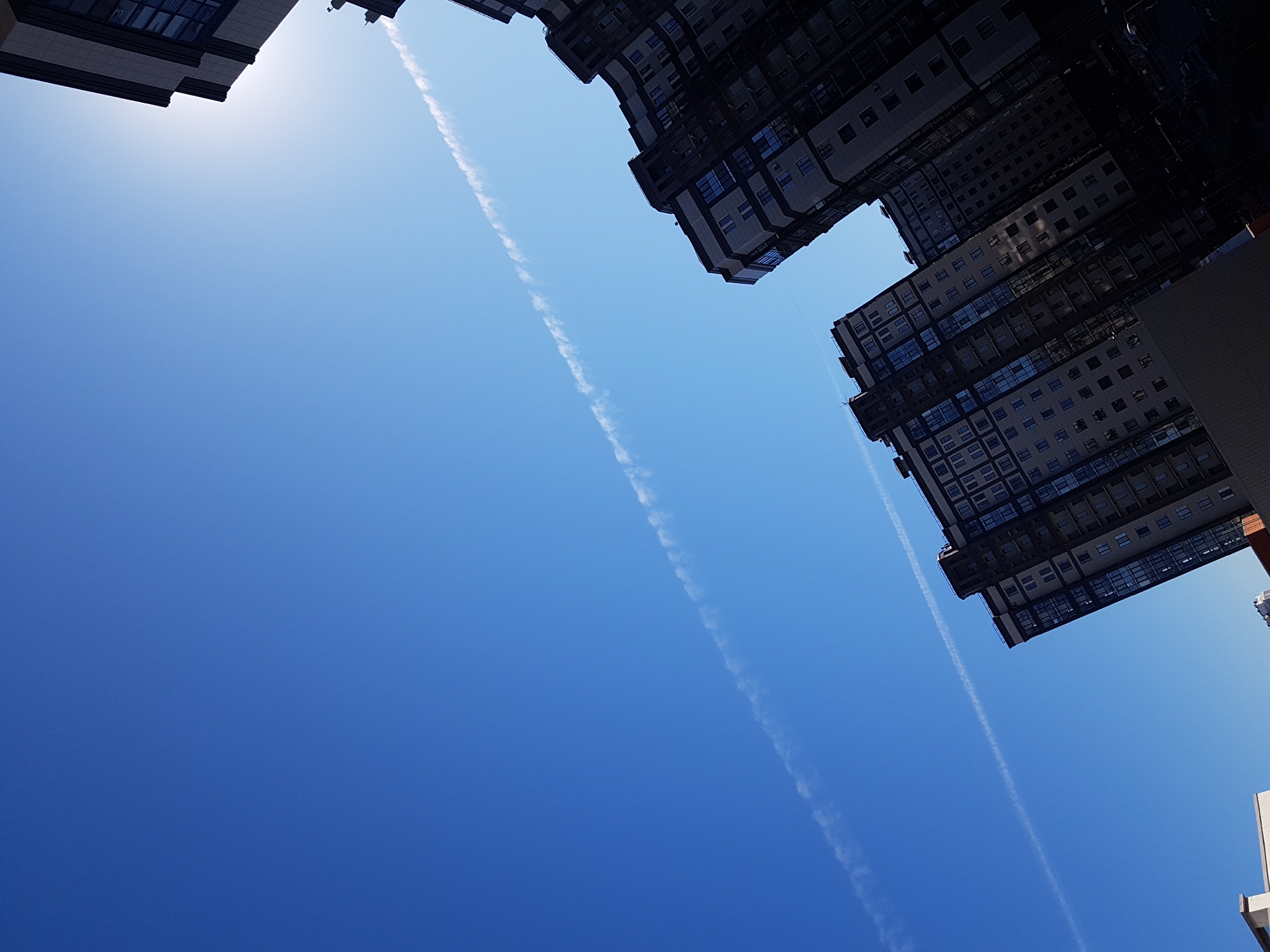 The image, taken from a low angle, features tall buildings against a clear blue sky in Saint Petersburg, Russia. Two contrails cut diagonally across the sky. The buildings are densely constructed, with numerous windows visible. The sunlight suggests a daytime scene.