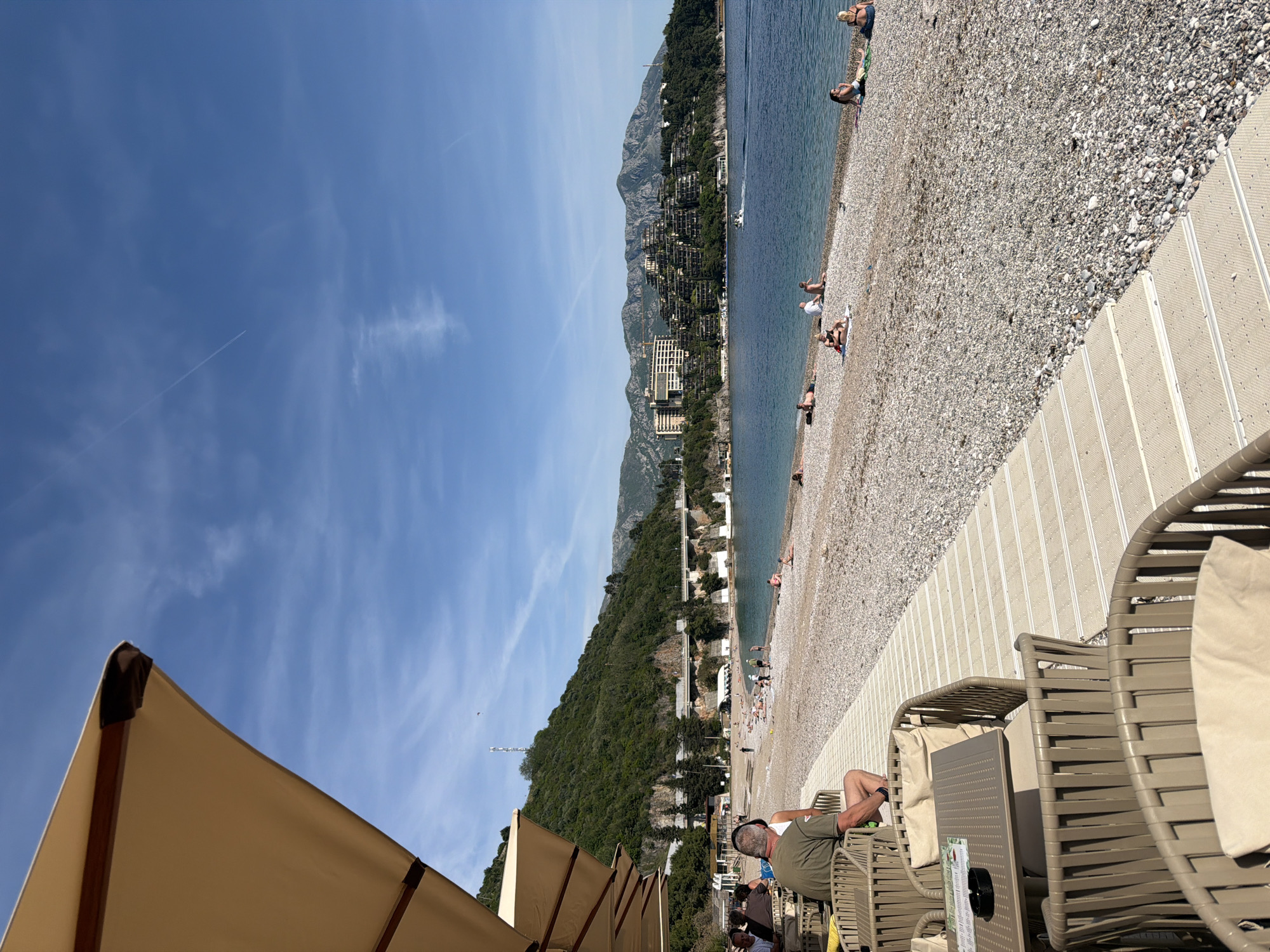 The image captures a sunny day at a pebble beach in Budva, Montenegro. The scene is oriented with the coastline running diagonally from the upper right to the lower left.
In the foreground, there's a patio area with lounge chairs and beige umbrellas. A man wearing a green shirt and headphones is relaxing in one of the chairs. There are tables and other chairs nearby.
Beyond the patio, the beach stretches along the Adriatic Sea. People are sunbathing and relaxing on the pebble beach. A small boat is visible in the water. The shoreline is lined with buildings, including hotels and apartments.
In the background, mountains rise up behind the city and the coastline. The sky is blue with wispy clouds.