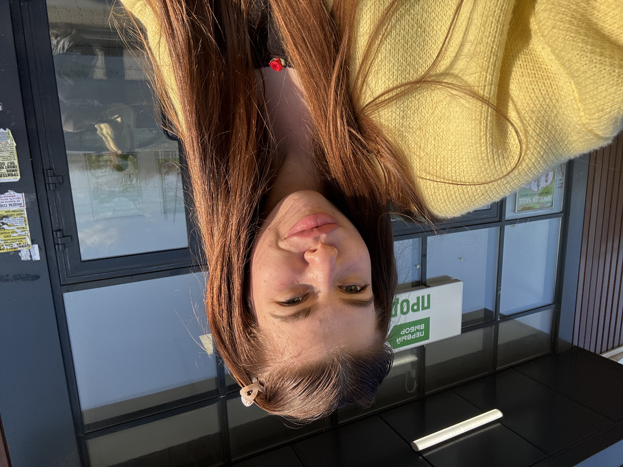Here is a detailed description of the image:
**Scene:** The photo shows a woman with long brown hair and a yellow sweater. The photo is oriented upside down. She is standing in front of a store in the city of Речицы, Russia.
**People:** The only person is a woman with fair skin, long brown hair, and pink lips. She is wearing a yellow sweater and has a small hair clip in her hair.
**Objects:** The photo features a store window with some advertisements and notices posted on it. There are reflections in the windows. A sign says "ПЕРВЫЙ ВЫБОР ПРОД" in Russian. Also seen are strips of light.
**Actions:** The woman is standing still, looking at the camera, and tilting her head.
**Environment:** The background is an urban setting, likely a street in front of a commercial building. The lighting appears to be daytime and slightly sunny.