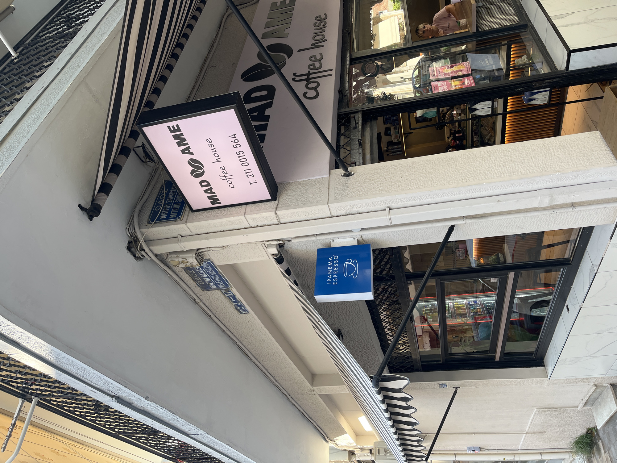 This image captures the exterior of a coffee shop in Athens, Greece, specifically on the street labeled "ODOS MELETIOU VASILEIOU" and indicated by house numbers "3-27". The building has a modern facade with a prominent awning featuring black and white stripes. Two signs are visible: one in pink light displaying "MAD AME coffee house" and a phone number "T. 211 0015 564," and another smaller blue sign that reads "IPANEMA ESPRESSO". The coffee shop has large windows, through which the interior is partially visible. Inside, shelves stocked with products and a counter are evident. A woman with blonde hair, wearing a pink shirt, is seated at a table inside the shop, looking towards the right. The street appears to be a narrow urban setting.