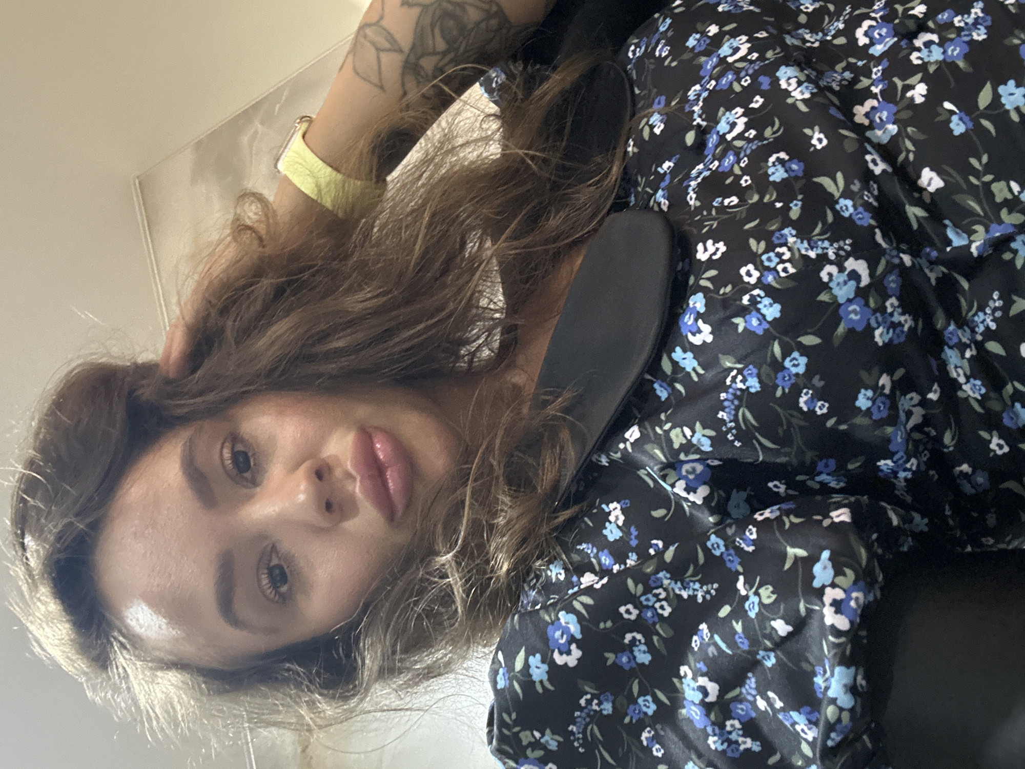 The image is a close-up selfie of a woman. She has long, brown, wavy hair, with some strands falling forward and obscuring parts of her face. Her head is tilted slightly upwards and to her right. She is wearing a black dress with a small floral print in shades of blue, white, and green. A black collar or lapel is visible at the neckline of the dress. On her left wrist, she wears a bright yellow smartwatch. Her right hand is raised and touching her hair near the crown of her head. The background appears to be an indoor setting with a textured, possibly wallpapered wall and a hint of a light fixture. There are no visible elements that would suggest a specific location within Duvankul', Russia.